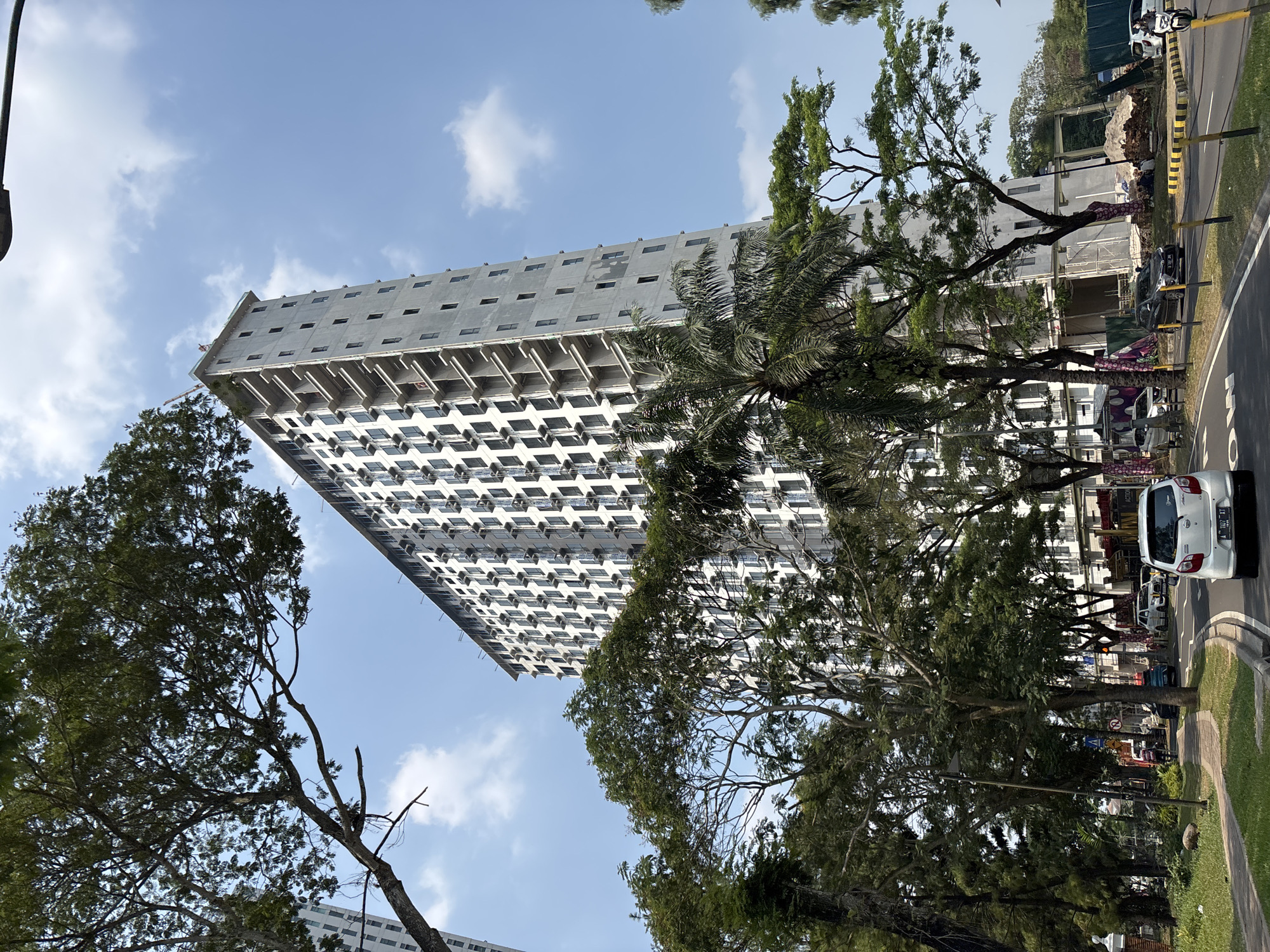 The image captures a view of a tall, unfinished apartment building under construction in Bencongan Indah, Indonesia. The building is a prominent feature, stretching high into the clear blue sky with scattered white clouds. Its exterior is concrete, with numerous small, dark windows visible. Balconies are in various stages of completion, some appearing open and others enclosed. Construction equipment and materials, including scaffolding and piles of dirt, are visible around the base of the building, indicating ongoing work.
In the foreground, a street curves through the scene, with several cars driving on it. A white car is positioned in the lower right quadrant of the image, facing away from the viewer. Trees, characterized by their lush green foliage and sprawling branches, flank the street and partially obscure the view of the building. A palm tree is also present, adding to the tropical feel of the environment.
On the left side of the street, a sidewalk is visible, bordered by grass and smaller plants. A lamppost stands on the sidewalk. The overall impression is that of an urban developing area, where modern construction is interspersed with established greenery. There are a few people scattered around the construction site, some sitting near the base of the building, appearing to be workers or observers. The lighting suggests it is daytime, likely late morning or early afternoon, given the brightness and clear skies.