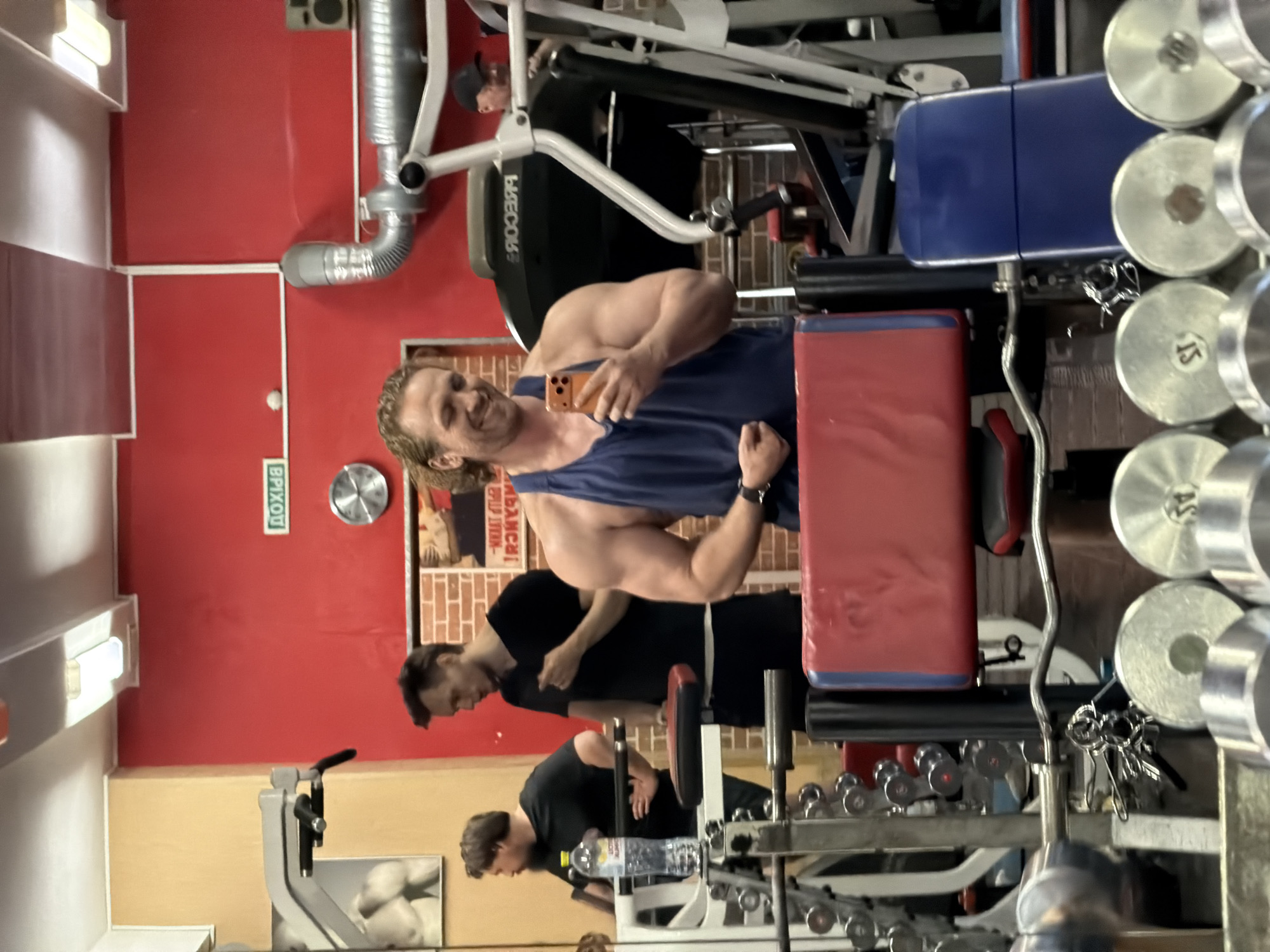 The image displays an indoor gym environment, specifically a fitness center in Saint Petersburg, Russia, identifiable by the visible Russian text. The central figure is a muscular man with long, wavy hair, wearing a blue tank top. He is smiling and taking a mirror selfie with an orange-cased smartphone while flexing his right bicep.
Several other individuals are present and visible primarily through reflections. Another man in a black t-shirt is seen in the reflection behind the main subject. Further back, a third man, also in a black t-shirt, appears to be working out, with a water bottle placed on a machine nearby. A fourth person, wearing a black cap, is partially visible in the background reflection near large gym equipment.
The gym features striking red walls with white trim. A green sign with the Russian word "ВЫХОД" (EXIT) is prominently displayed on a red wall. Gym equipment includes various dumbbells, with at least one marked "15," and large exercise machines, one of which bears the "PRECOR" brand name. A set of keys hangs near some weights. A poster with Russian text, possibly reading "МЫ - ЧЕМПИОНЫ!" (WE ARE CHAMPIONS!), is visible on a section of the wall designed to resemble red brick. The overall atmosphere appears bright and active, with the main subject conveying a sense of happiness and confidence.