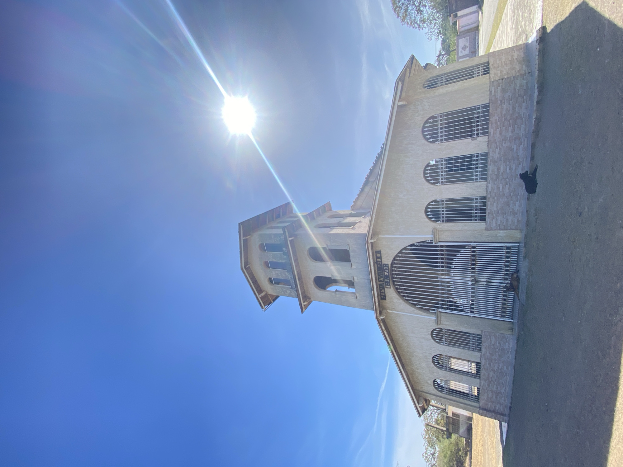 The image shows a light-colored building with a tower in Juan Griego, Venezuela. The architecture includes arched windows and a large arched gate or entrance with bars. The tower has a similar arched design. The structure appears to be a church. The sky is bright blue with the sun prominently positioned. A dark object, possibly a dog, is near the building's base. The surrounding area includes grass and some distant trees.