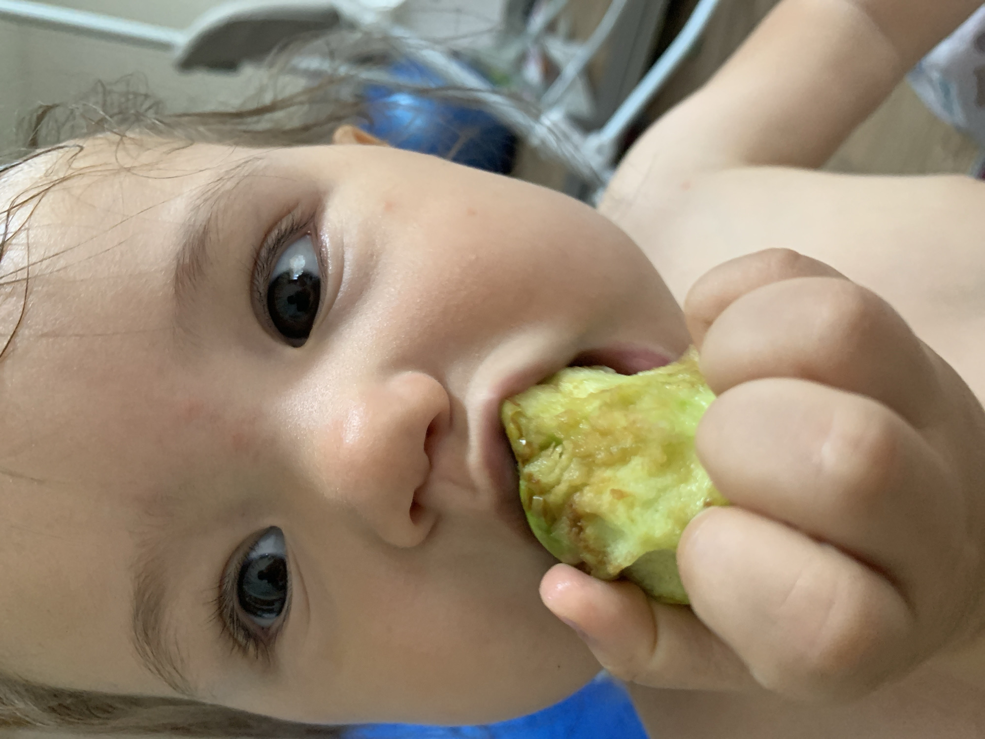 A close-up, eye-level, medium shot captures a young child's face as they eat a piece of green food, possibly avocado. The child's left eye is in focus, looking to the right of the frame. Their mouth is open, biting into the food. The child's left hand is visible, holding the food. The background is blurred and appears to be an indoor setting with hints of blue and white objects. The image does not provide any information or context regarding the city of Chaikovsky, Russia.