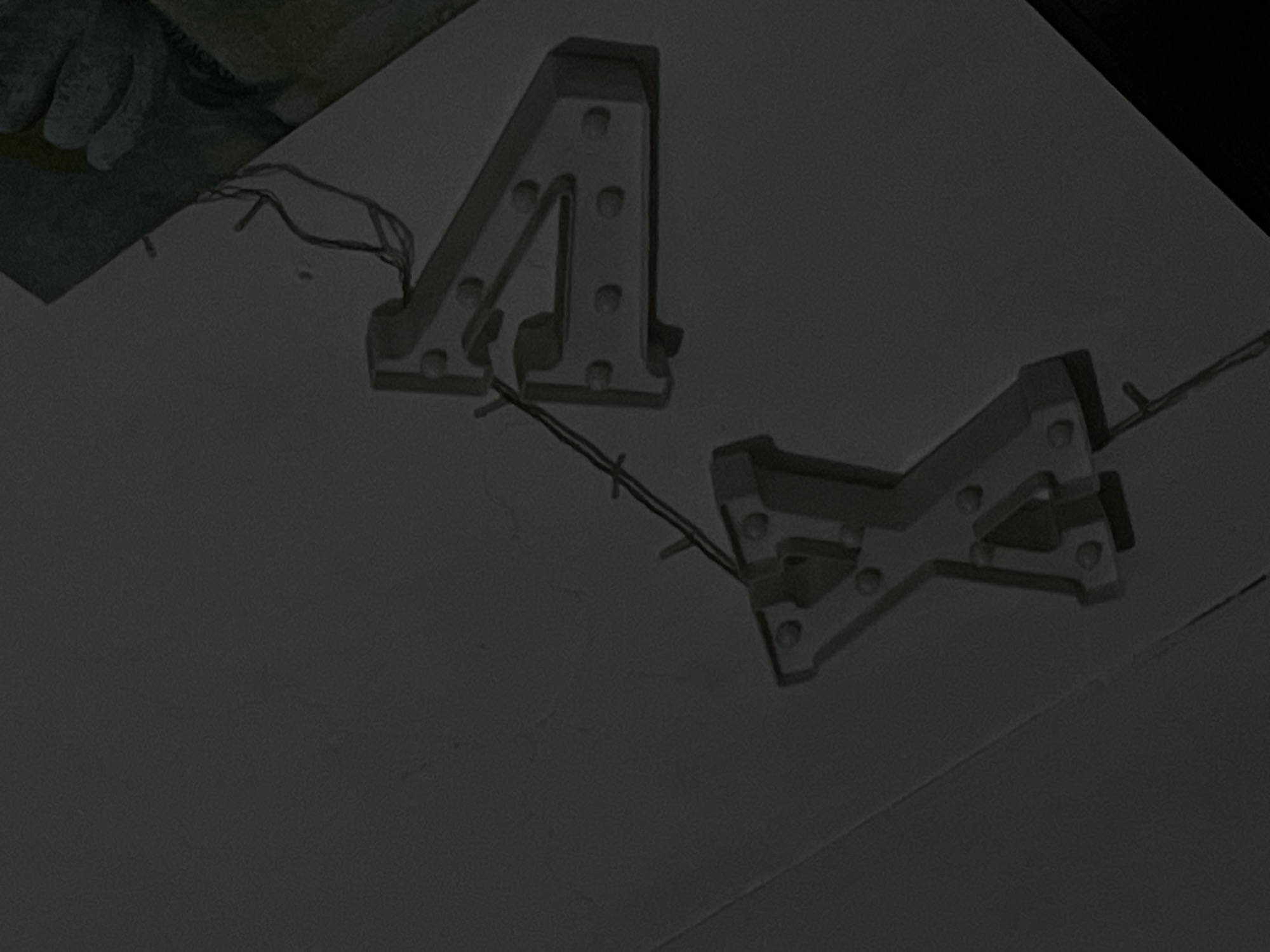 The image shows two illuminated marquee letters, an "A" and an "X", against a dark, neutral background. The letters are white and appear to be made of plastic with bulbs set into them. The "A" is larger and positioned to the left, while the "X" is smaller and to the right, slightly overlapping the "A". Both letters have exposed wiring, suggesting they are either in the process of being set up or have been disassembled. The lighting is dim, creating shadows that emphasize the three-dimensional form of the letters. In the top left corner, a portion of a hand wearing a glove or sleeve is visible, adding a hint of human presence. The overall scene is static, with no obvious activity. The context is unclear, but the presence of marquee letters might suggest a celebration or event. The lack of ambient light suggests an indoor setting or nighttime.