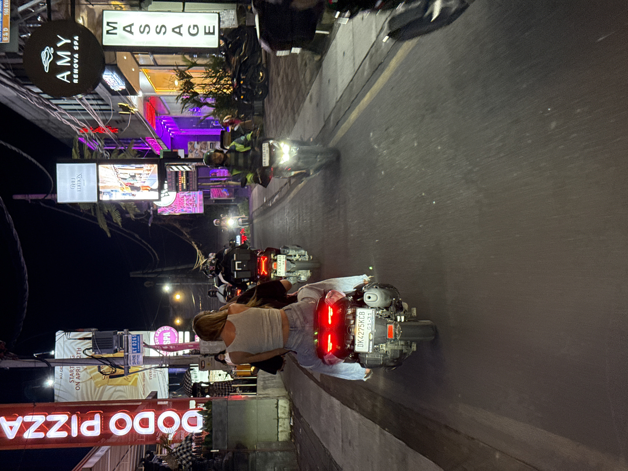 The image captures a lively night scene on a commercial street in North Kuta, Bali, Indonesia, with the perspective tilted to showcase the street, scooters, and illuminated storefronts. Several people are visible, primarily riding scooters. In the foreground, a woman with blonde hair, wearing a light top and jeans, is a passenger on a scooter, facing away from the camera. Its red taillights are prominent, and the license plate "DK 4275 KCB" with "01-31" is visible. Further down the street, another individual on a scooter is seen wearing a helmet and a reflective vest, with bright headlights. A second scooter's license plate, "DK 3364 AER" with "02-30", is also discernible.
The street is brightly lit by numerous artificial light sources, including vibrant neon signs and illuminated lightboxes from various businesses. The overall atmosphere suggests a bustling evening in a popular area.
Visible text on signs includes: "MASSAGE", "AMY RENOVA SPA", "DODO PIZZA" (appearing upside down), "BALI EDITION", "FASHION DESIGN", "CUT CANGGU SPA", "HYDRAFACIA", "KU DE", "START G ON APRI 2TH", "B&D", and "160 KVA". Additional text "KT 0265" and "TARARA" are also present. The "DK" prefix on the license plates confirms the location in Bali.