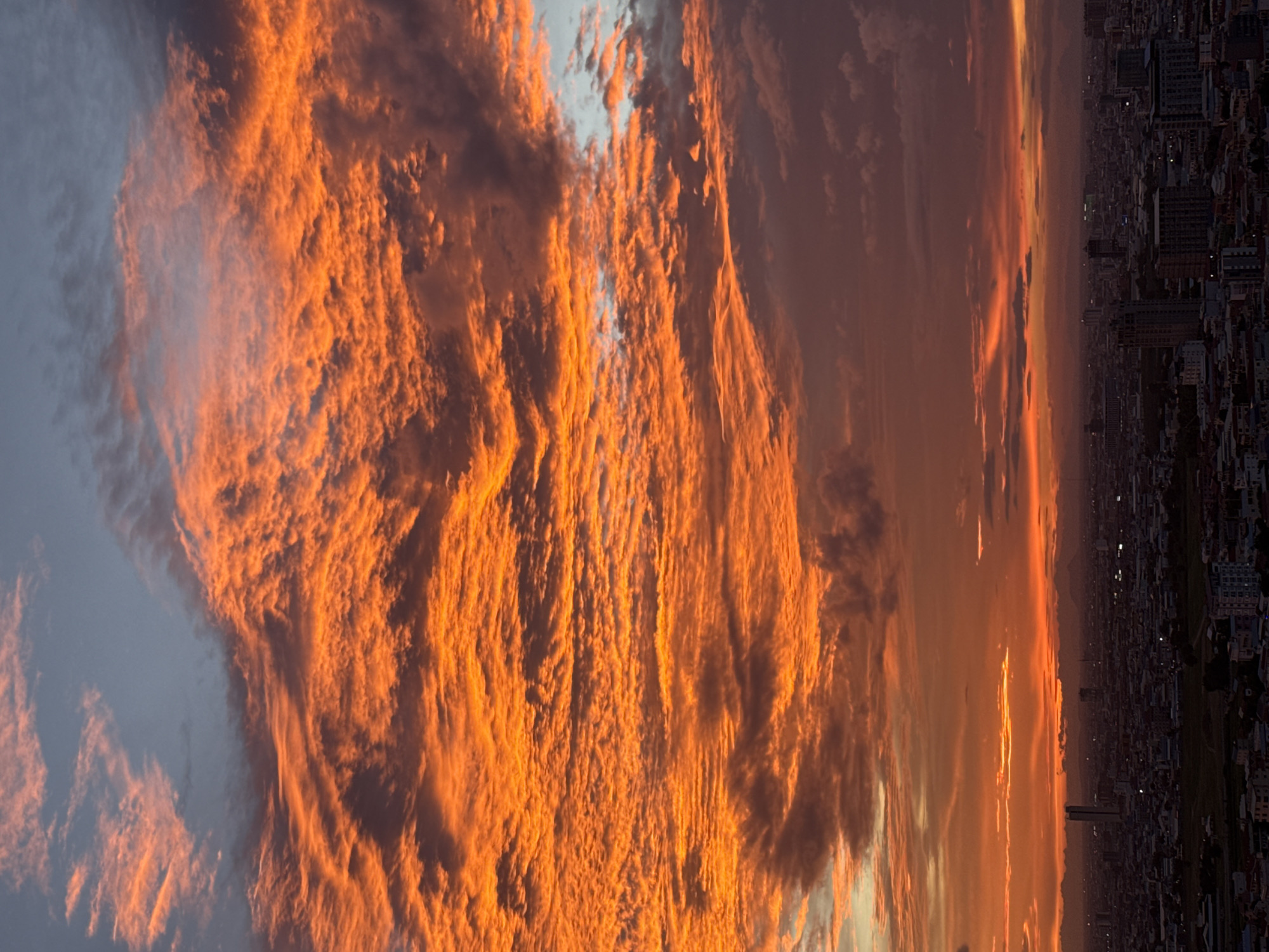 This image captures a vibrant sunset over the city of Khan Mean Chey, Cambodia. The sky is dominated by fiery orange and red clouds, creating a dramatic and picturesque scene. Below the intensely colored sky, the city sprawls out, a dense collection of buildings and structures that are silhouetted against the warm glow. A few tall buildings stand out in the urban landscape. The overall impression is one of a breathtaking natural phenomenon occurring above a busy, developing cityscape.