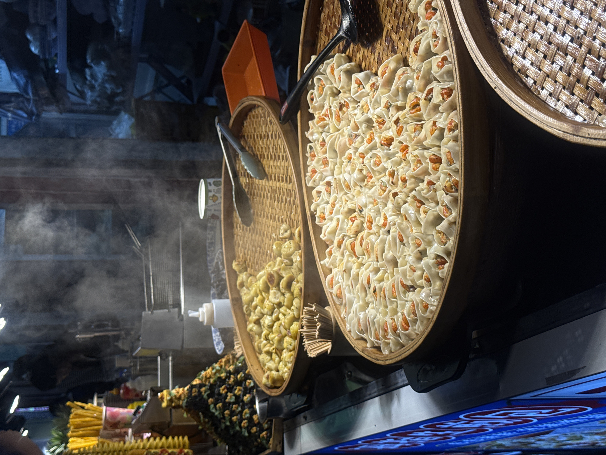 At a bustling food stall in Qinhuangdao, China, a vendor is preparing steamed dumplings. Two large bamboo steamers are filled with the dumplings, one with a golden, partially cooked batch and another with a finished batch of delicate, white dumplings filled with orange ingredients, likely shrimp. Steam rises from the dumplings, indicating they are freshly cooked. Tongs and a ladle rest on the steamers, ready for serving. In the background, shelves are stocked with various items, and other food items like yellow skewers are visible. The scene captures the vibrant atmosphere of a street food market, with the focus on the delicious and visually appealing dumplings.