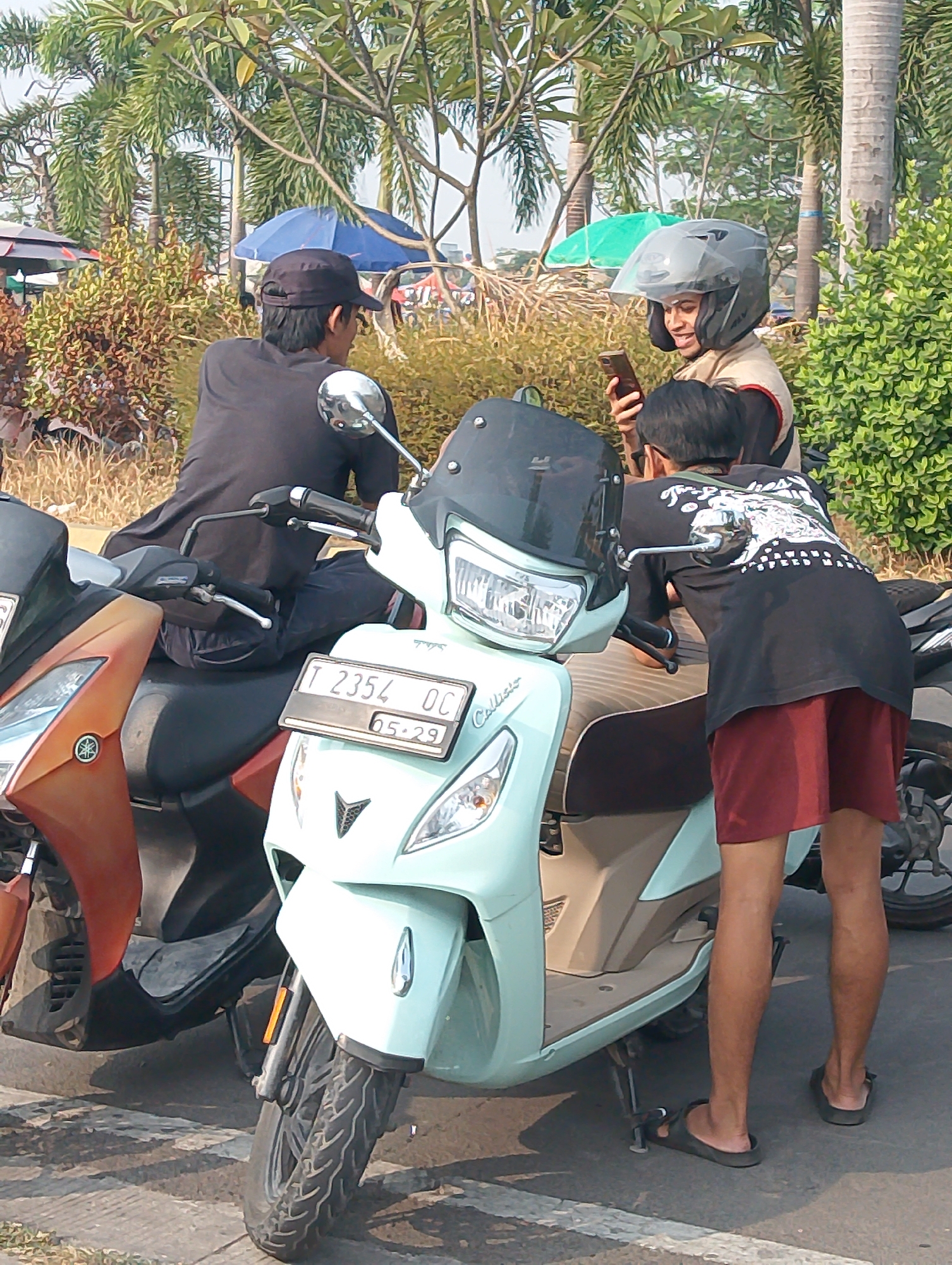 Three individuals, two adult males and one adult female, are positioned around two parked scooters outdoors in Karawang, Indonesia. The female, wearing a helmet and a light-colored shirt, is looking at her phone. One male, wearing a dark baseball cap and a black shirt, is seated on a scooter. The other male, wearing a black t-shirt with a graphic print and dark red shorts, stands near the front of a light blue scooter, his legs visible. The scooters appear to be parked on a paved surface, possibly a street or parking lot. Vegetation, including palm trees and bushes, is visible in the background. An assortment of umbrellas are also present in the background, suggesting a market or outdoor event. The lighting indicates it is daytime.