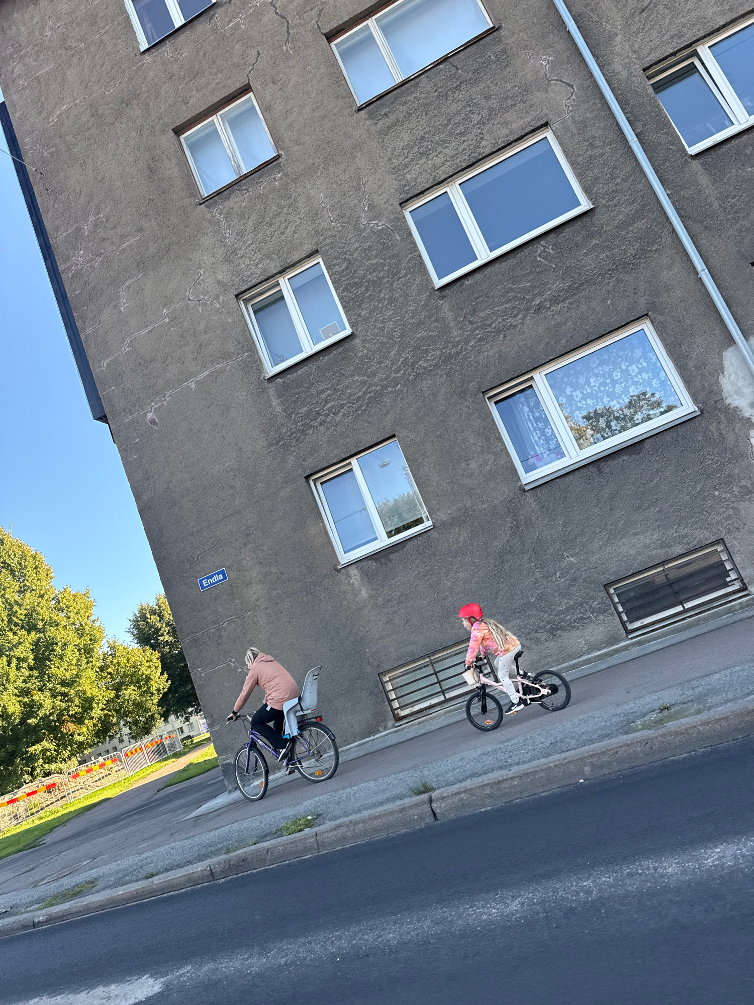 The image depicts two individuals cycling along a sidewalk in front of a weathered, grey apartment building. The building features multiple rectangular windows, some of which reflect the sky and surrounding greenery. A blue street sign with the inscription "Endla" is affixed to the building's facade. On the left side of the image, lush green trees are visible, and a section of a yellow and red striped barrier separates the sidewalk from a grassy area. The foreground shows a street with a curb. The perspective of the photograph is angled upwards, emphasizing the height of the building.
One cyclist, positioned further down the sidewalk, is an adult wearing a pink jacket and riding a dark-colored bicycle with a child seat. The child seat is empty. The other cyclist, closer to the foreground, is a child wearing a pink helmet and a colorful tie-dye jacket, riding a pink bicycle. Both cyclists are moving from left to right.
The scene is characteristic of an urban residential area. Based on the street sign "Endla" and the architectural style, the location is likely Endla Street in Tallinn, Estonia, a common residential area in the city. The image captures a casual, everyday moment of people enjoying outdoor activity in an urban setting.