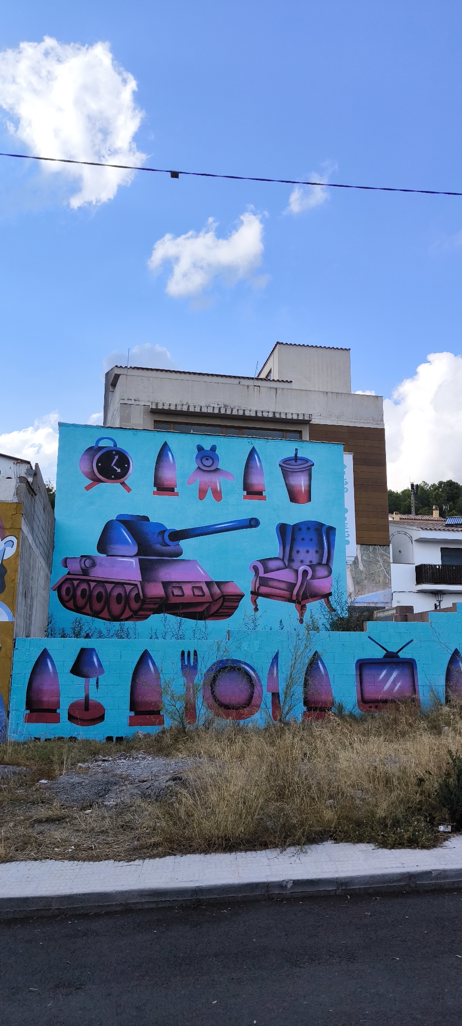 The image shows a vibrant mural painted on a rough concrete wall, likely located in Fanzara, Spain, a town known for its street art. The mural, set against a bright turquoise background, features a surreal juxtaposition of military hardware and everyday objects, all rendered in a bold, graphic style with gradient shading from purple to pink.
Dominating the center of the mural is a cartoonish, purple and pink tank with a long barrel. Above the tank, arranged in a row, are various objects: a vintage-style alarm clock with a black face and red numbers, a small, pink, teddy-bear-like figure, and two stylized ammunition rounds, one of which is a lighter purple with a red base. To the right of the tank, a plush armchair, also rendered in shades of purple and pink, appears to float. Another ammunition round is depicted to the right of the armchair.
Below the tank, another row of objects is painted. This includes a desk lamp with a blue shade and red base, more ammunition rounds, a fork, a plate, and a knife. To the far right of this lower row, a vintage television set with a screen displaying static is visible.
The mural is situated outdoors, with dry, yellowing grass and weeds growing in front of the wall, suggesting an arid or unkempt environment. A paved sidewalk and a dark asphalt road are visible in the foreground. In the background, parts of buildings can be seen, including a light-colored structure with brown wood paneling and a white building with red tile roofs, indicative of a Mediterranean architectural style. The sky above is a bright blue, dotted with fluffy white clouds. A dark power line with a small, black object suspended from it stretches across the upper portion of the image. The mural appears to be unsigned and the overall impression is one of playful subversion, merging symbols of conflict with domesticity.