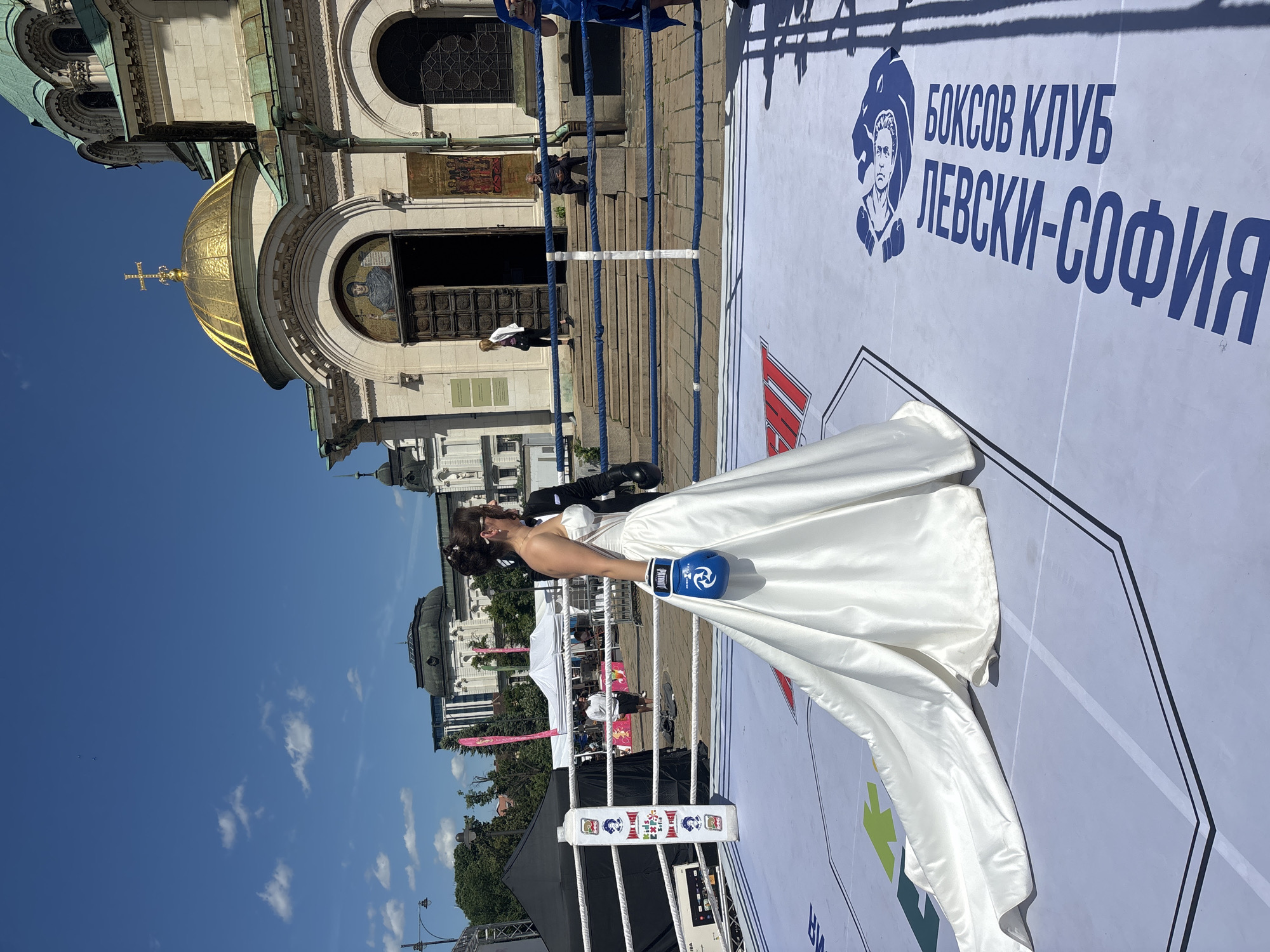 The image depicts a woman in a white wedding dress and blue boxing gloves standing inside a boxing ring. In the background, the Alexander Nevsky Cathedral in Sofia, Bulgaria, is visible. The boxing ring has ropes and corner posts, and there is signage on the floor with the words "БОКСОВ КЛУБ ЛЕВСКИ-СОФИЯ" (Boxing Club Levski-Sofia) along with a logo. The sky is blue with some clouds.