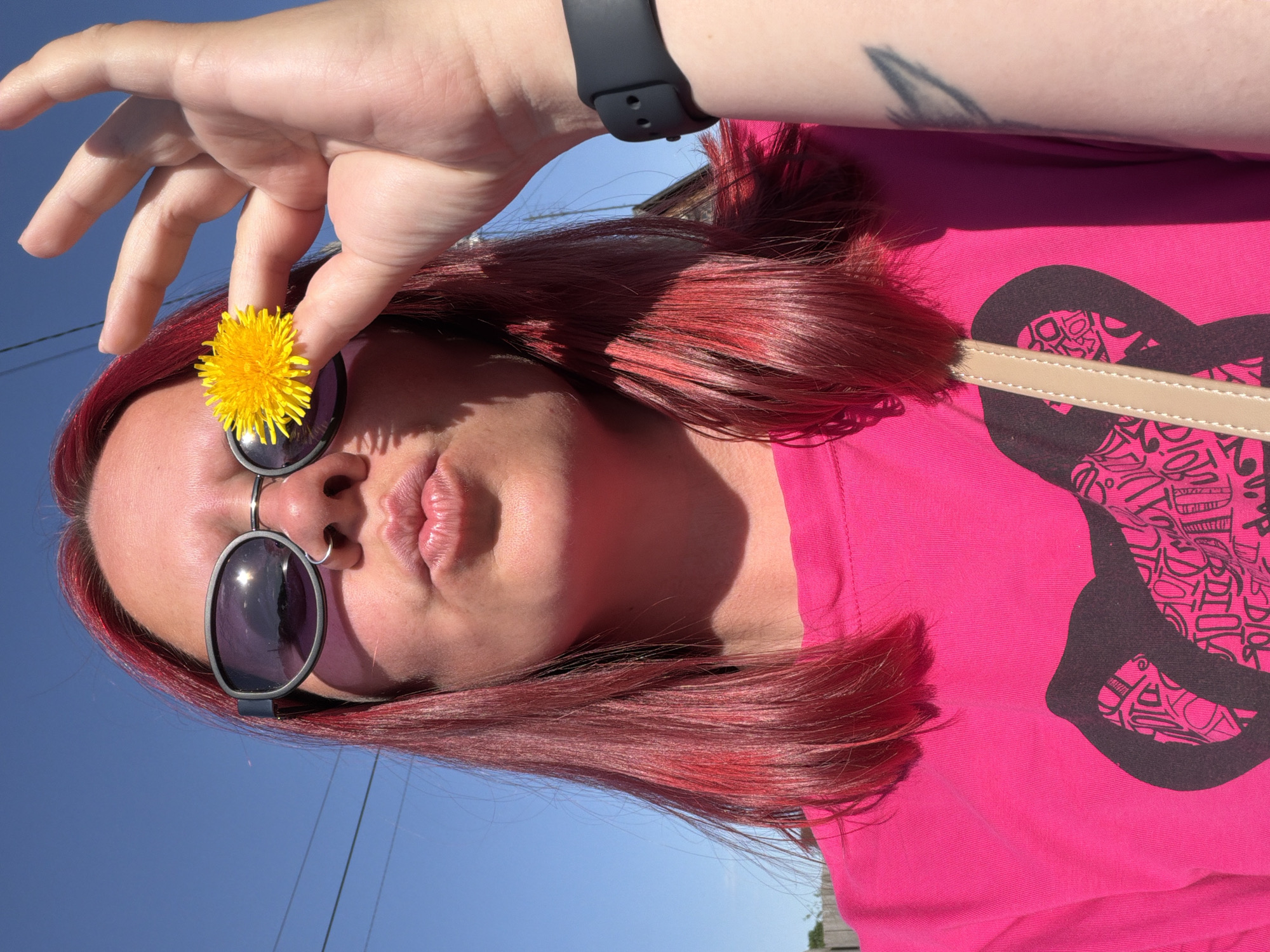 A fair-skinned person with red hair is posing for a selfie outdoors in Chaikovsky, Russia. The subject is wearing sunglasses with dark lenses and a pink T-shirt with a black graphic design. A black smartwatch is visible on their left wrist. A dandelion flower is being held in front of one eye. In the background, there is a clear blue sky, power lines, and some buildings.