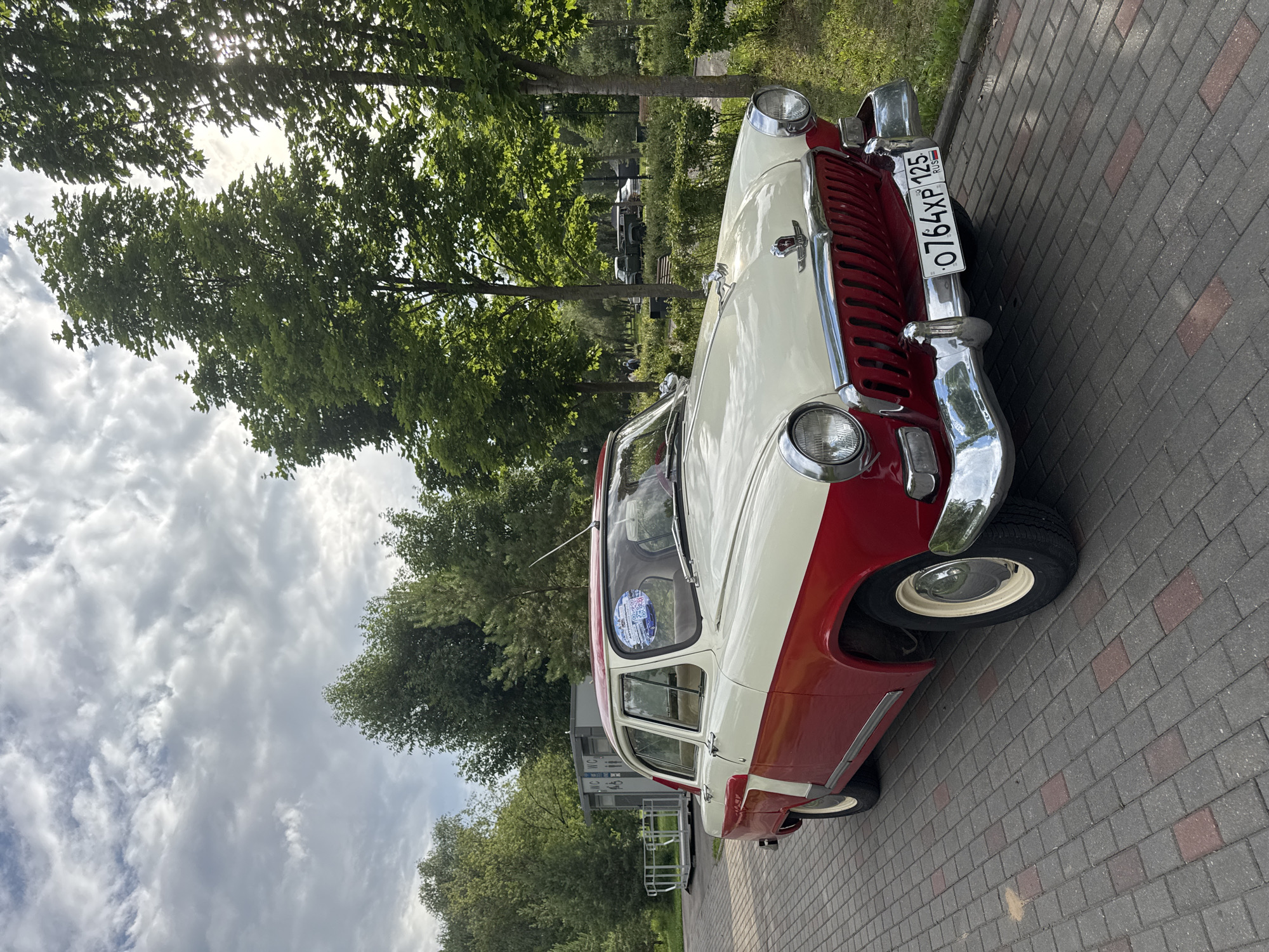 A vintage, two-tone red and white car, possibly a GAZ-21 Volga, is parked on a brick walkway. The car's chrome grille and headlights gleam. A license plate with the text "0764XP 125 RUS" is visible on the front bumper. The car is positioned at an angle, with its front facing towards the right side of the frame.
The setting appears to be outdoors, with lush green trees and foliage in the background. The sky is partly cloudy, with sunlight filtering through the leaves of the trees, suggesting it is daytime. In the distance, other vehicles and a building can be seen. The overall mood is calm and serene. The image was taken in Moscow, Russia. There are no people clearly visible in the frame.