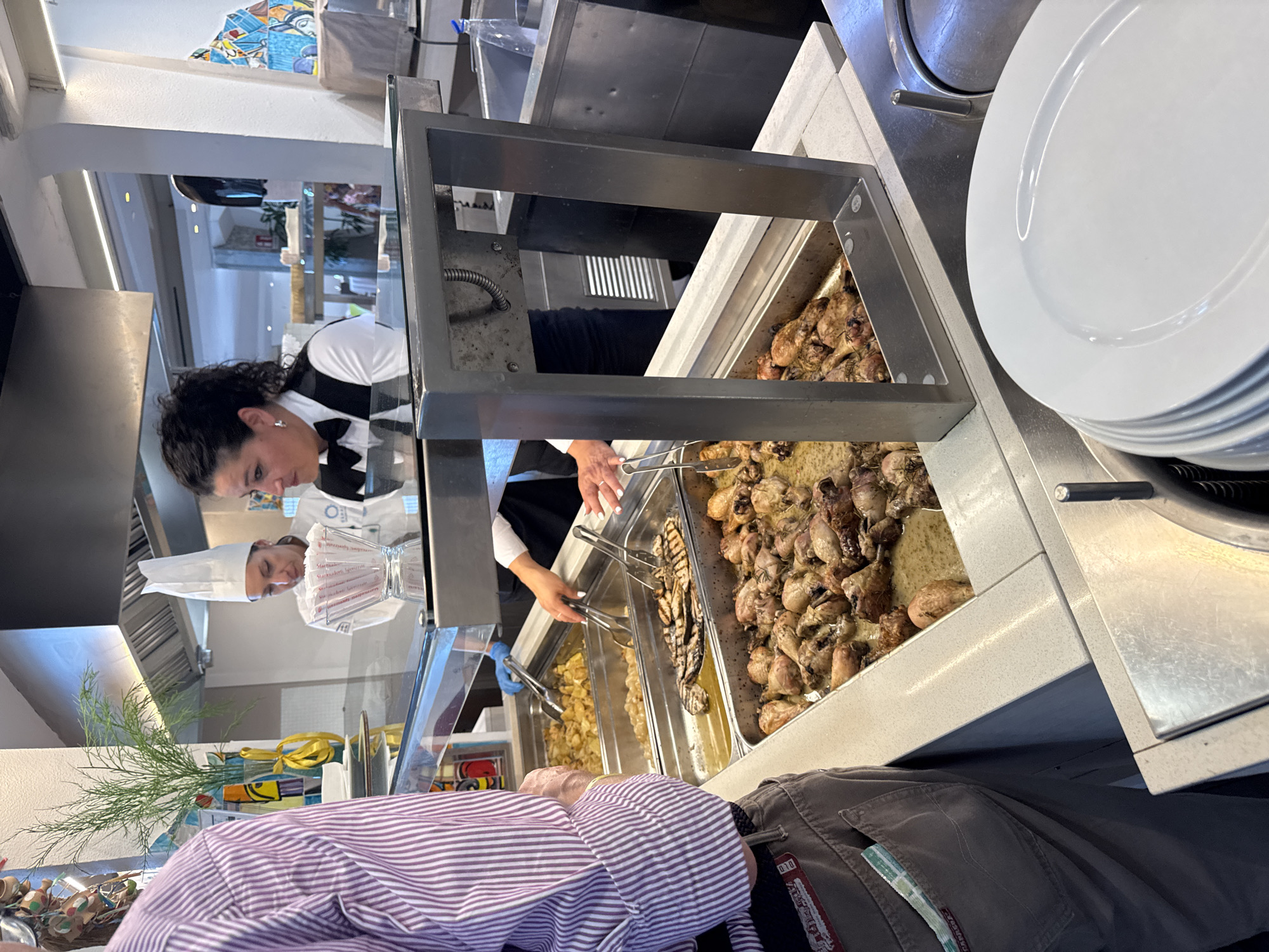 A person in a purple and white striped shirt is standing in line at a buffet. They are facing away from the camera. To their right, there are several food serving trays. One tray contains what appears to be cooked chicken legs with a sauce. Another tray holds grilled items, possibly fish or meat. A third tray contains a golden-colored dish, likely a side like potatoes or rice. In the background, two people are visible. One person, wearing a white chef's hat and a white shirt, is looking down at a menu or recipe card. Another person, wearing a black vest over a white shirt, is also looking in the same direction. A stack of white plates is visible to the right of the food trays. The scene appears to be indoors, possibly a restaurant or catering establishment. The overall impression is that of a busy food service setting. The food looks appetizing, and the people appear engaged in their tasks. There is no indication of the time of day or weather conditions. The location is Isola delle Femmine, Italy.