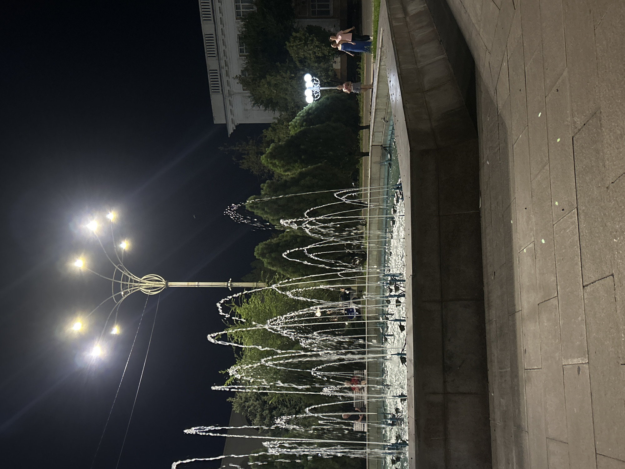 The image depicts a nighttime scene in front of the Taraz city administration building in Taraz, Kazakhstan. A large, multi-tiered fountain is the central focus, with numerous water jets spraying upwards, illuminated by the ambient light. The ground in the foreground is paved with large, rectangular stones. In the mid-ground, a distinct streetlamp with a unique, branched design is visible, its bright lights cutting through the darkness. Behind the fountain, lush green trees are interspersed with the architecture of the city administration building, which is illuminated and shows classical design elements, including columns and a prominent cornice. Two individuals are walking away from the camera on a paved pathway to the right of the fountain, one wearing a blue dress and the other a pink top and dark pants. On a bench to the left, partially obscured by the fountain's water, two people are seated. The overall atmosphere is one of a well-lit public space at night.