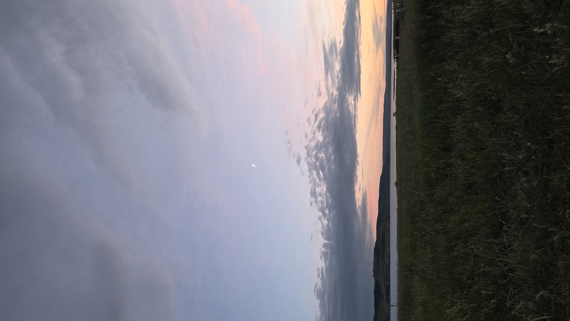 The image depicts a serene landscape at dusk, likely in or near Kupoyarovo, Russia. In the foreground, a dense field of tall green grass stretches across the frame, with some stalks catching the fading light. Beyond the field, a body of water, possibly a river or lake, is visible, reflecting the sky. The horizon is marked by a line of rolling hills or a distant shoreline, dotted with indistinct structures and trees, suggesting a populated area. The sky above is a canvas of soft colors, transitioning from a pale blue at the top to hues of pink and orange near the horizon, indicative of sunset. Scattered across the sky are numerous clouds, some appearing dark and heavy, while others are wispy and illuminated by the last rays of the sun. A small, crescent moon is faintly visible in the upper portion of the sky. A few small figures, appearing to be people, are discernible on the distant shoreline to the right, suggesting a recreational area. The overall atmosphere is tranquil and evocative of a peaceful evening in a natural setting.
