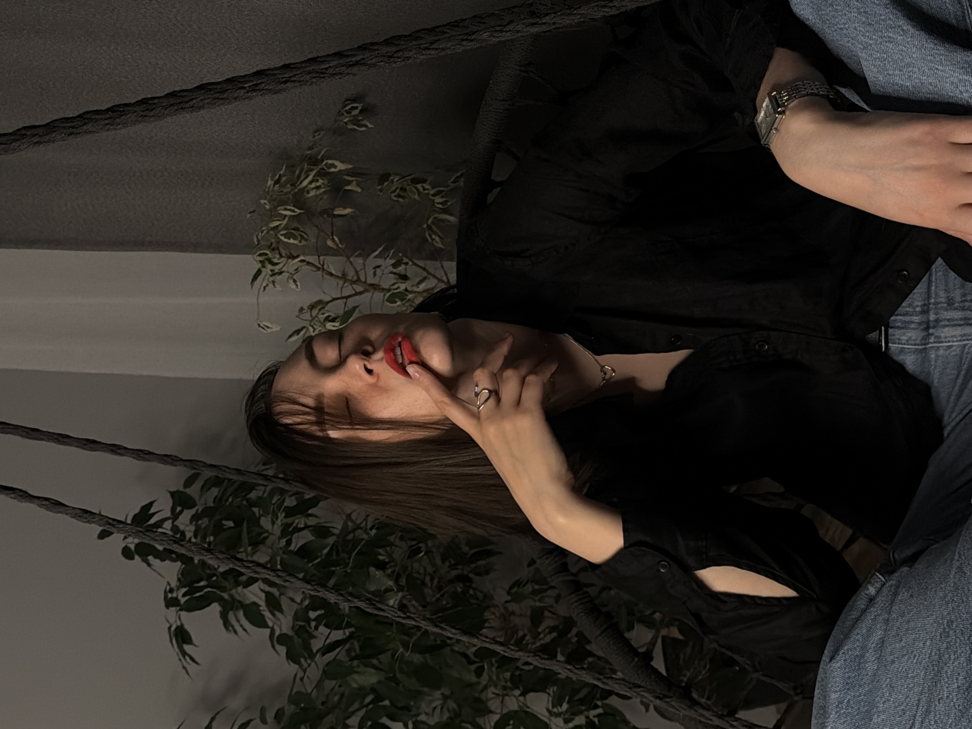 The image shows a young woman lying down in an indoor setting. She has dark hair, is wearing a black collared shirt or jacket, and blue denim pants are visible on the right side of the frame. On her left wrist, she wears a metallic watch with a rectangular face. Her right hand is raised to her face, with a ring visible on her index finger, and her finger gently touches her chin or mouth. Her eyes are closed or nearly closed, and she has bright red lipstick.
Two thick, dark ropes diagonally traverse the upper and mid-sections of the frame, suggesting she might be in a swing or hammock. Lush green plants with distinctive variegated leaves are scattered in the background and foreground, adding natural elements to the scene. The lighting is dim and diffused, contributing to a calm and contemplative atmosphere, indicative of evening or nighttime. The woman's pose and closed eyes suggest relaxation or introspection. There is no visible text in the image. The setting is likely a cozy interior in Chaikovsky, Russia.