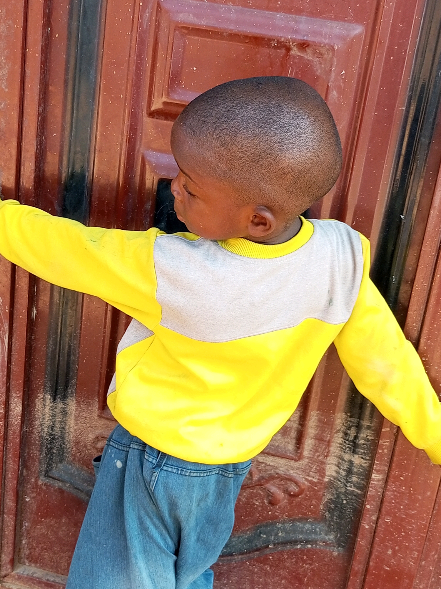 A young child, viewed from the side and slightly from behind, stands with their back to a dark red, paneled door. The child has dark skin, a short haircut, and is wearing a bright yellow long-sleeved shirt with a grey horizontal band across the chest and shoulders, and blue denim jeans. The child's left arm is extended outwards, with their hand out of frame. The door has vertical panels with some textured details and appears to be made of metal. The lighting suggests an outdoor setting, possibly in the city of Tempure, Nigeria.
