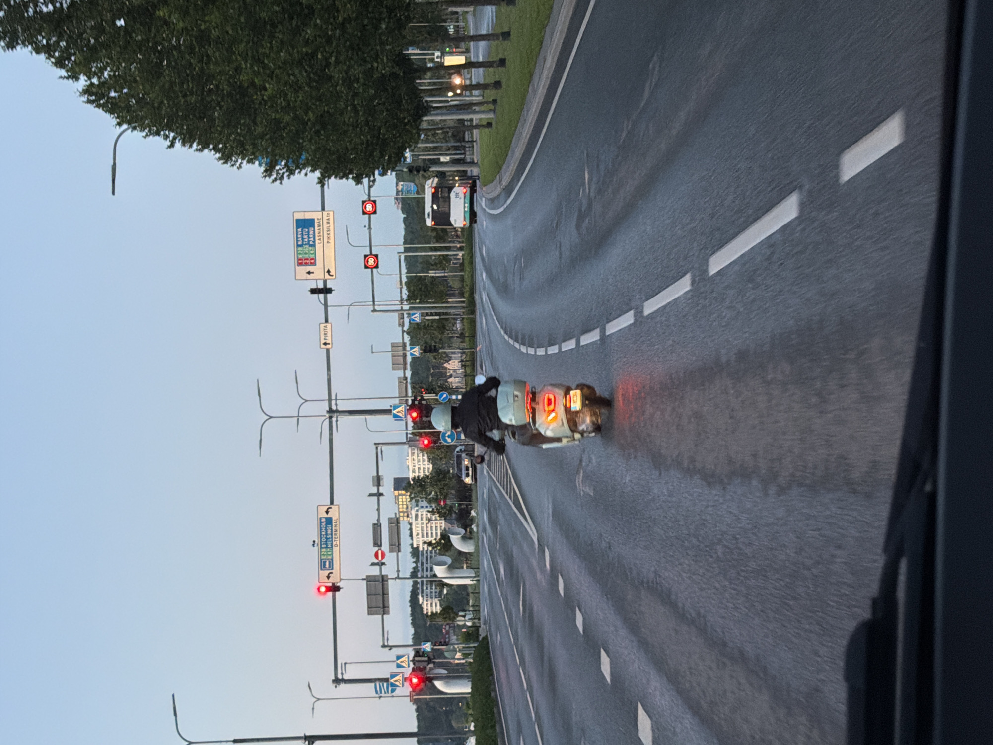 The image captures a scene on a wet road in Tallinn, Estonia, likely during dusk or early morning due to the lighting conditions. A person is riding a light-colored scooter on the right lane of the multi-lane road. The scooter rider is wearing a dark jacket and a light-colored helmet. The scooter's brake lights are illuminated red. In the distance, a white bus with green accents is visible, traveling in the same direction. Traffic lights are red at an intersection ahead, indicating that vehicles are stopped. Road signs indicate directions to various locations, including "D-TERMINAL," "STOCKHOLM," and "HELSINGI" on one sign, and "NARVA," "TARTU," "PARNU," "LASNAMÄE," and "PIKKSI LMA 1b" on another. The road surface is asphalt and appears damp, reflecting the ambient light. Trees line the right side of the road, and modern buildings are visible in the background. The perspective suggests the image is taken from a vehicle.
