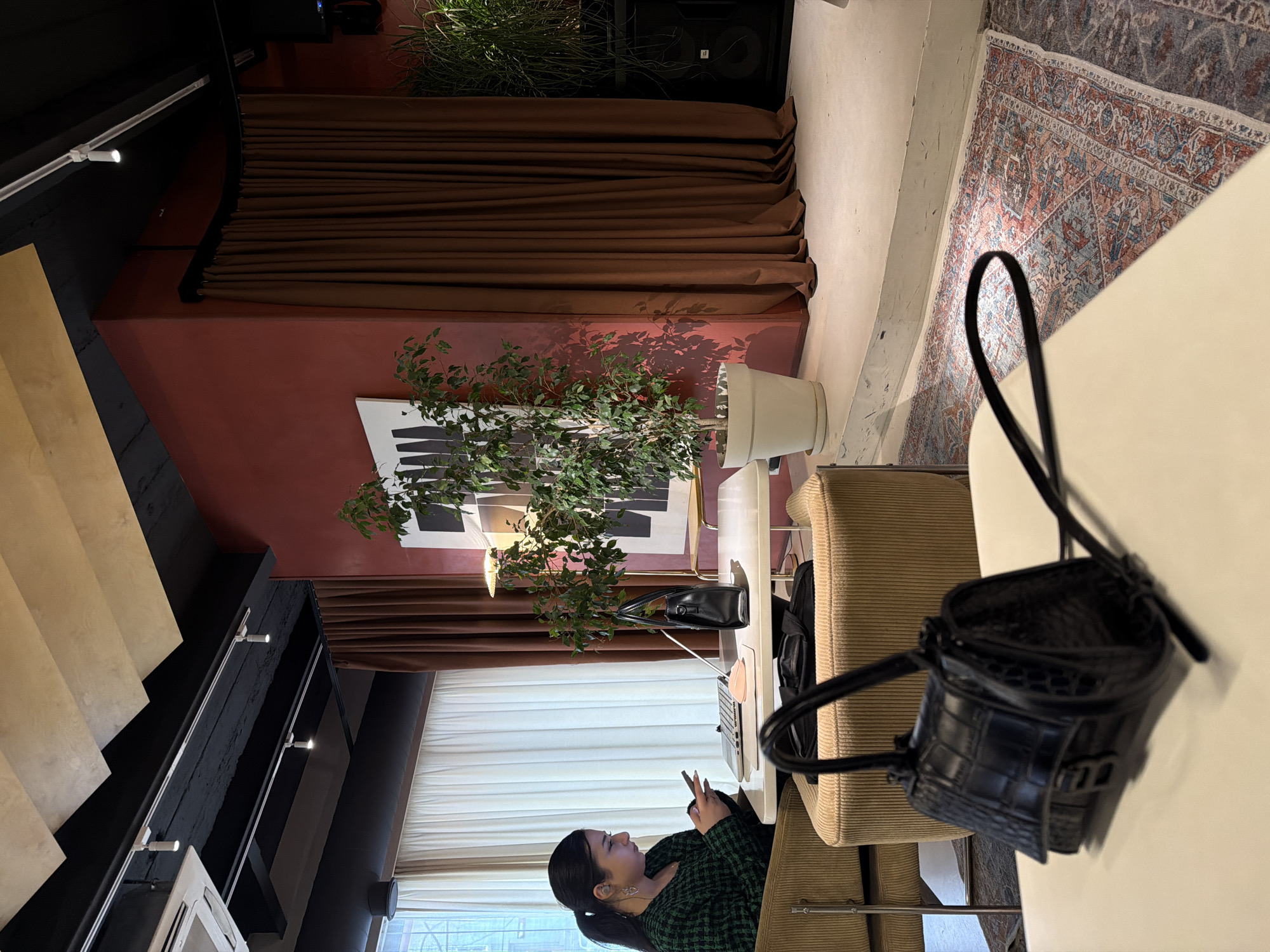 An indoor scene within a commercial establishment in Yerevan, Armenia, is captured from a slightly elevated perspective. A female with dark hair, pulled back into a ponytail, is seated in a beige ribbed armchair on the left mid-ground. She wears a dark green textured top and is actively viewing and manipulating a smartphone with both hands. In front of her, on a white rectangular table, an open silver laptop and a small pink object are positioned. A black, structured handbag with two top handles rests near the laptop.
To the right of the seated female, a large potted plant with green foliage stands in a white planter on the floor. Behind this plant, a dark red wall displays a white rectangular art piece featuring vertical black markings. The red wall extends towards a floor-length brown curtain on the right, which conceals a portion of a raised platform. Large black speaker units are partially visible on this platform, adjacent to another potted plant.
In the immediate foreground, a small black handbag with a crocodile-embossed texture and a prominent "B" shaped buckle rests on a white tabletop. Below this table, a patterned rug displaying red, blue, and beige motifs covers a section of the floor. The ceiling above is black, featuring exposed beams, white track lighting fixtures, and beige geometric sound-dampening panels. A white air conditioning unit is mounted on the ceiling in the upper left corner. A window covered by sheer white curtains is visible on the left, through which blurred exterior elements are discernible.