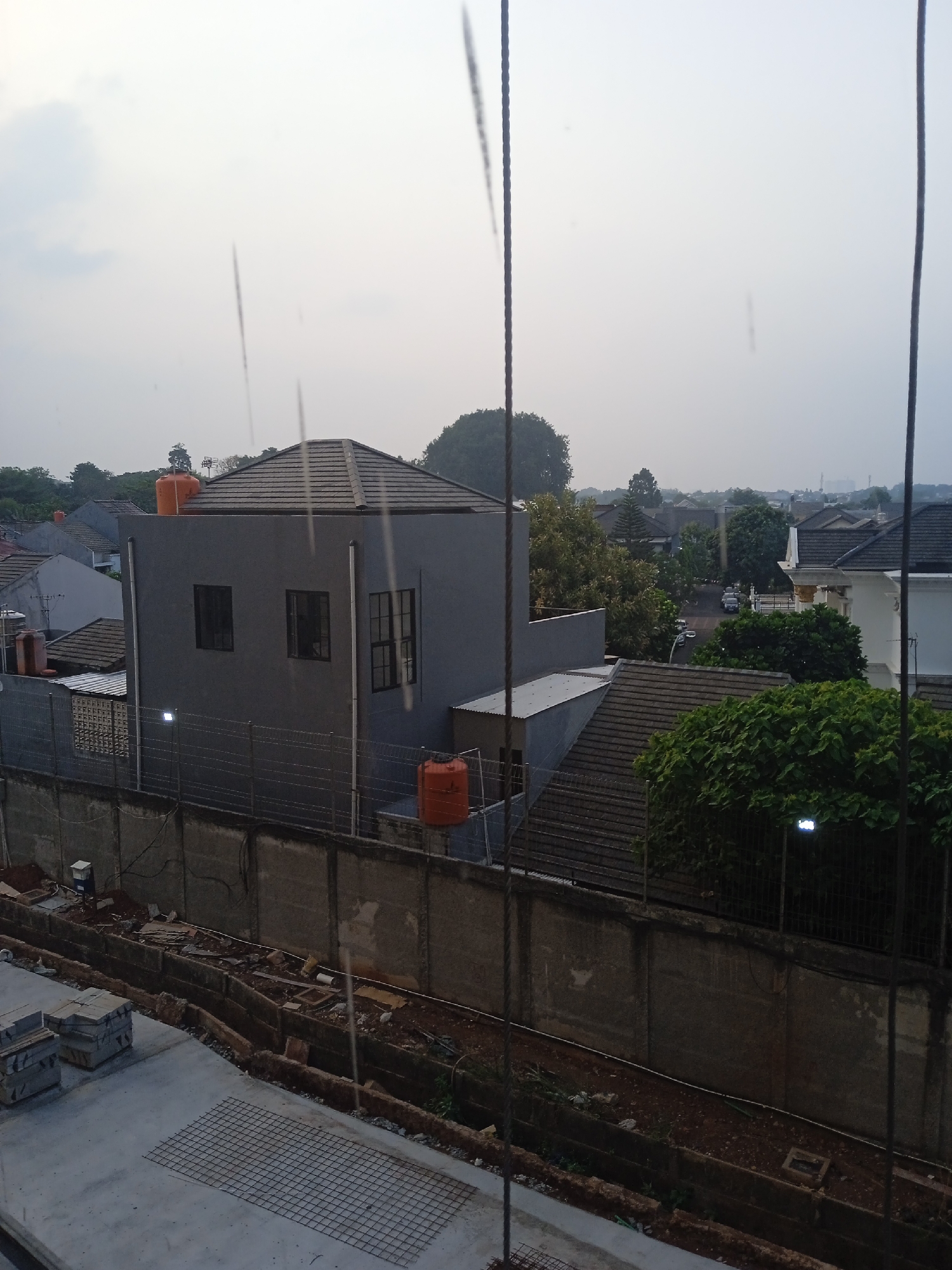 The image captures an outdoor scene in the city of Bekasi, Indonesia, likely during daylight hours with overcast skies. In the foreground, a construction site is visible, with stacks of grey building materials and a metal mesh grid laid out on a concrete surface. A trench runs along the edge of the construction area, filled with loose soil and some debris.
Further back, a concrete wall separates the construction site from a residential area. Beyond the wall, several houses are visible, characterized by their grey and white facades and dark tiled roofs. One prominent grey house features dark-framed windows and an orange water tank on its roof. Another orange water tank is situated near the wall on the right side of the image.
The background shows a street with a few parked cars, surrounded by lush green trees and more houses. The overall atmosphere suggests a suburban or developing urban environment in Bekasi. The image is partially obscured by vertical black lines, which appear to be part of the viewpoint or framing.