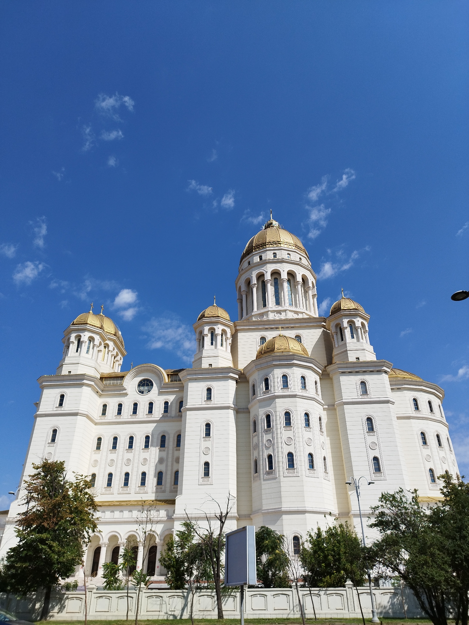 The image captures a low-angle, full-shot view of the Romanian Orthodox Patriarchal Cathedral in Bucharest, Romania, on a clear, sunny day with scattered white clouds. The large, white cathedral features multiple golden domes and numerous windows. The architecture is grand and ornate, with layered tiers and decorative elements. Trees and landscaping are visible in the foreground, bordering a white, decorative fence. A lamppost and a sign are also present. The sky is a vibrant blue, dominating the upper half of the image.