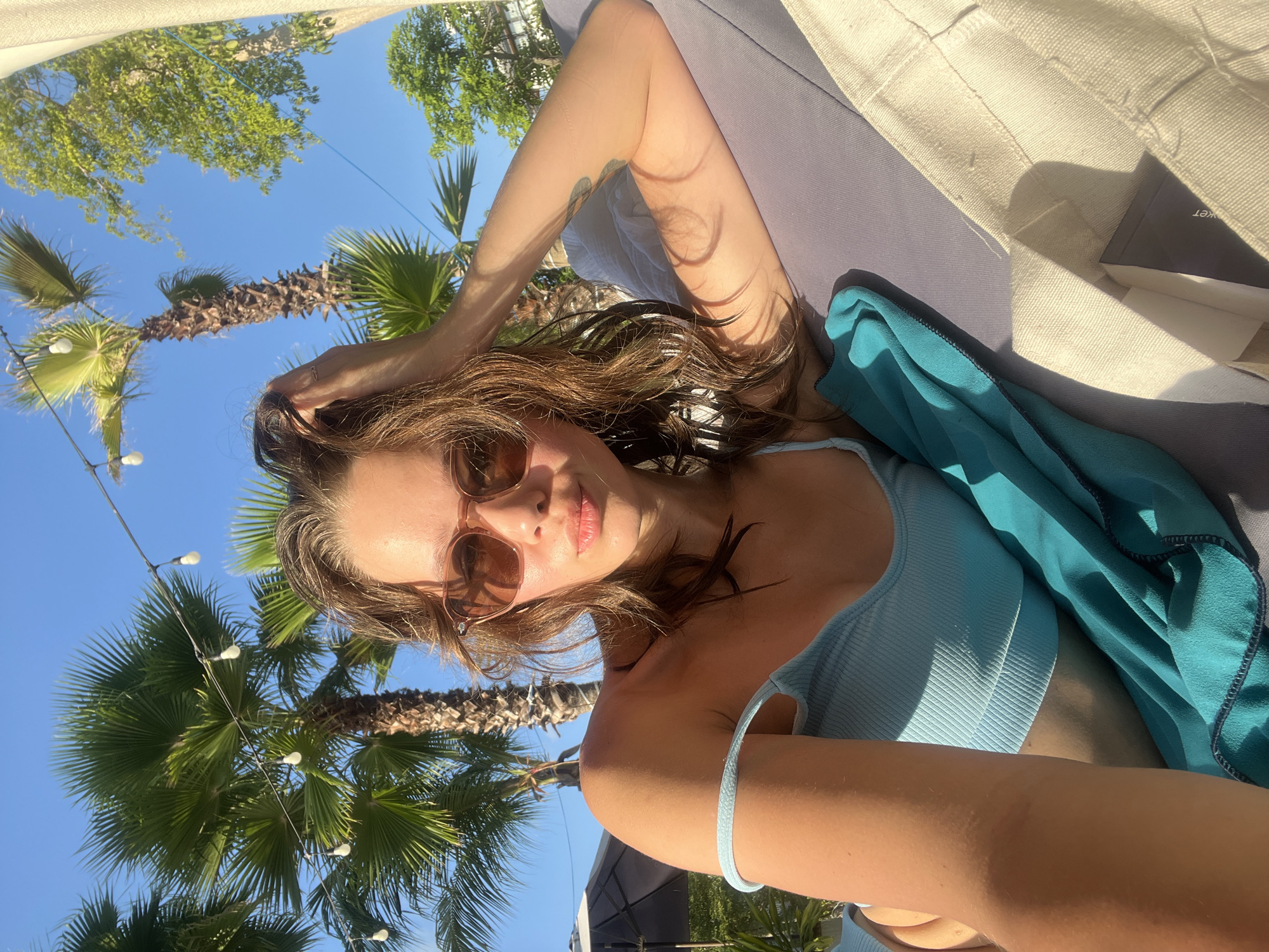 A woman with brown hair and sunglasses is lying on a lounge chair. She is wearing a light blue top and has a turquoise towel draped over her. Palm trees and string lights are visible in the background against a bright blue sky. The location is suggested as Nam Nha Trang Ward, Vietnam.