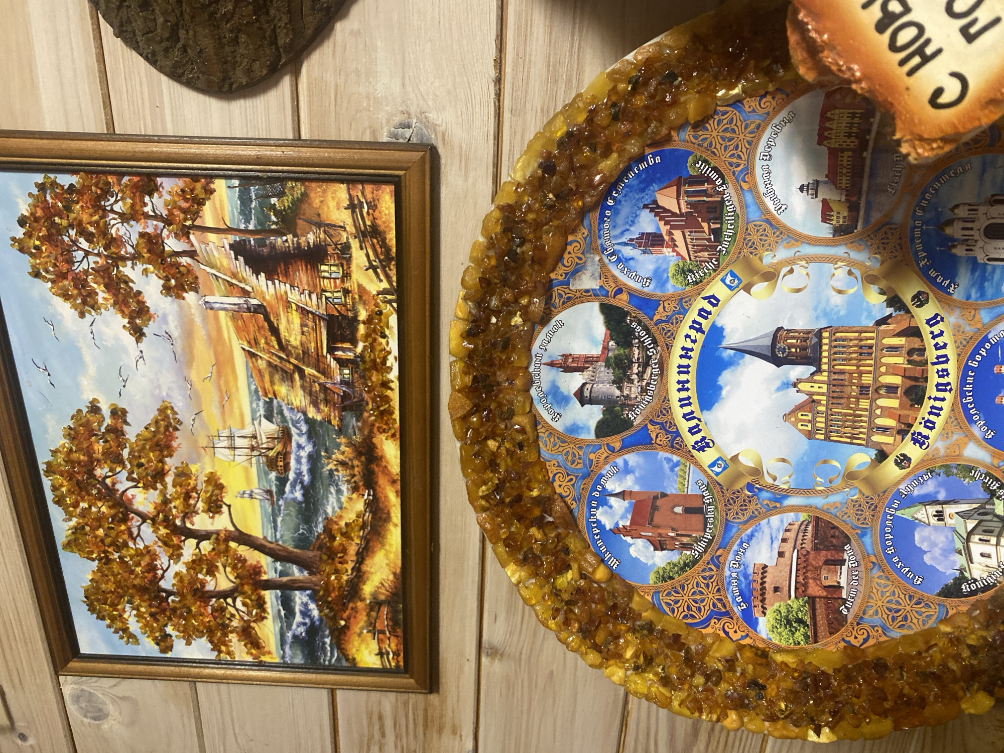 A framed painting of a seaside cottage with autumn trees and sailing ships hangs above an amber-encrusted decorative plate. The plate features circular images of historical buildings and landmarks, with Russian text labeling them. The background is a wooden wall. The image does not depict the city of Mitino, Russia.