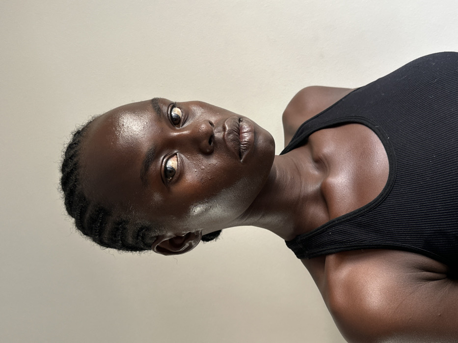 The image is a medium close-up portrait of an individual with dark skin, appearing to be a young adult female, positioned against a plain, light-toned background. Her hair is styled in tightly braided cornrows, pulled back from her forehead. She possesses prominent cheekbones, full lips, a straight nose, and wide-set eyes with light-colored irises, maintaining a direct gaze towards the viewer. The skin exhibits a natural sheen, particularly across the forehead, nose, and cheekbones. She is wearing a black, ribbed, racerback-style tank top, visible across her shoulders and upper chest. The background is a uniform, untextured light beige or off-white surface, providing no specific environmental details or indicators to determine a location within the city of Paris, France, or any other geographical area.