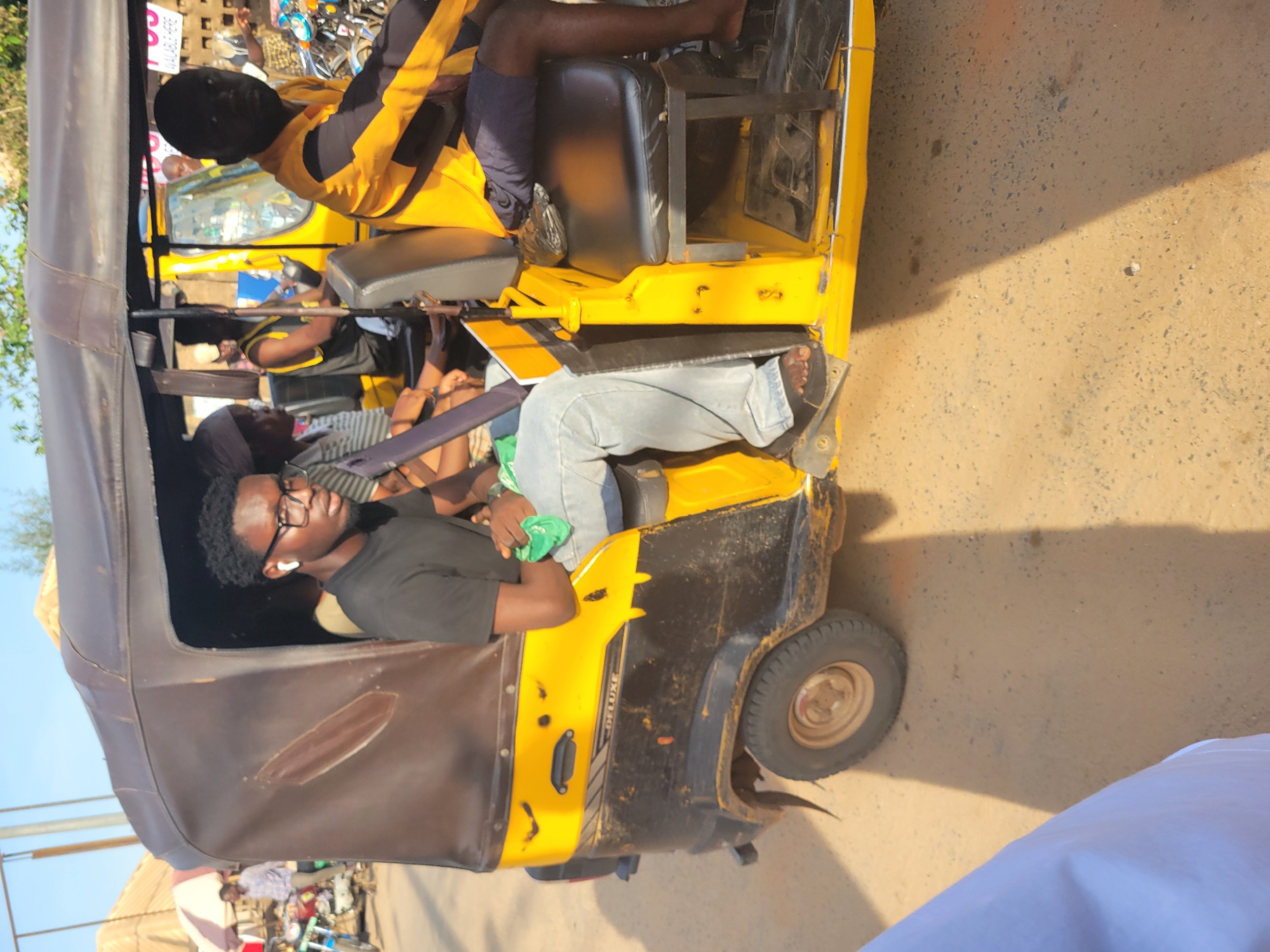In Egbagu, Nigeria, a yellow and black auto-rickshaw, commonly known as a "Keke Napep" or "Marwa", is parked on a dirt road. The auto-rickshaw appears to be carrying passengers. The driver, wearing a yellow and black long-sleeved shirt, is seated in the front right seat. A young man with glasses and white earbuds is in the passenger seat behind the driver, holding a green cloth. Another person, partially visible, is seated behind him. The auto-rickshaw has a brown canvas top and a black interior. The vehicle is weathered, with visible dirt and scuff marks on its exterior. The surrounding environment is dusty with some vegetation and buildings in the background. The sky is clear and blue, indicating a sunny day.