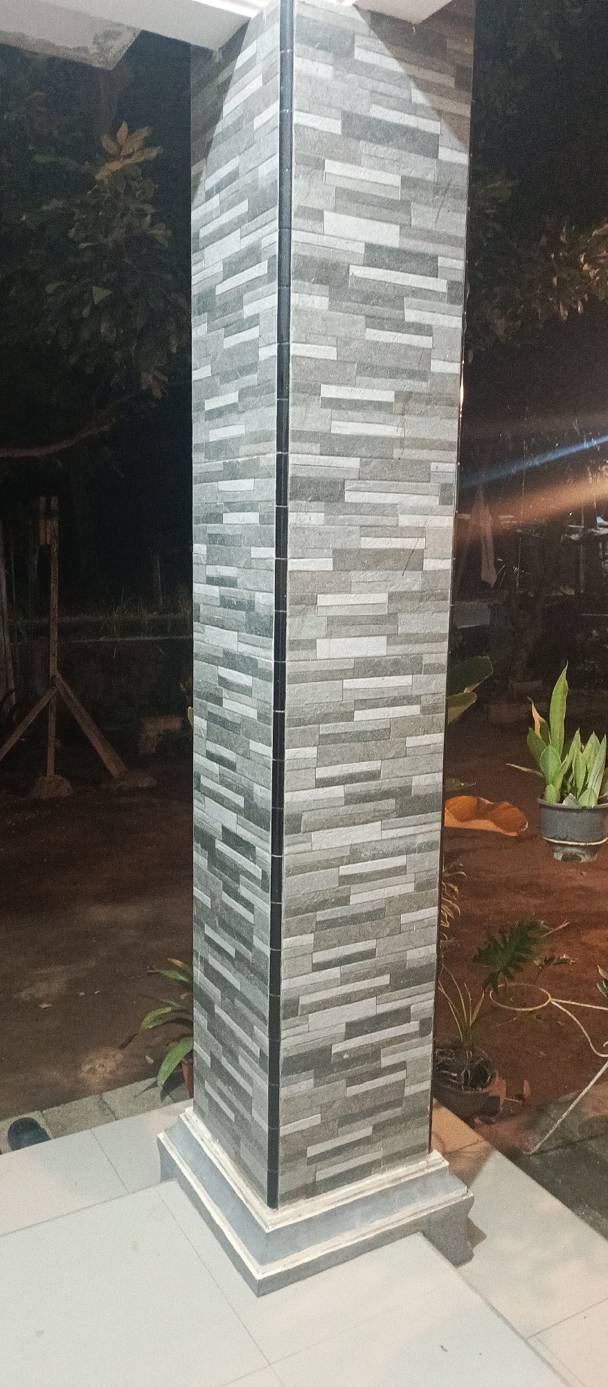 The image shows a close-up of a tiled pillar at night. The pillar is square and covered in rectangular tiles of varying shades of grey and white, arranged in a staggered pattern. There is a dark vertical seam running down the center of each visible face of the pillar. The pillar sits on a decorative base that is also grey and has multiple layers. The foreground shows a light-colored tiled surface, likely a floor or walkway. In the background, there are dark, out-of-focus areas with some foliage visible on the left. On the right side of the pillar, a potted plant with green leaves is illuminated, and there is a suggestion of other plants and outdoor elements. The lighting appears to be artificial, casting shadows and highlighting the textures of the pillar. The location is identified as Unknown, Unknown.
