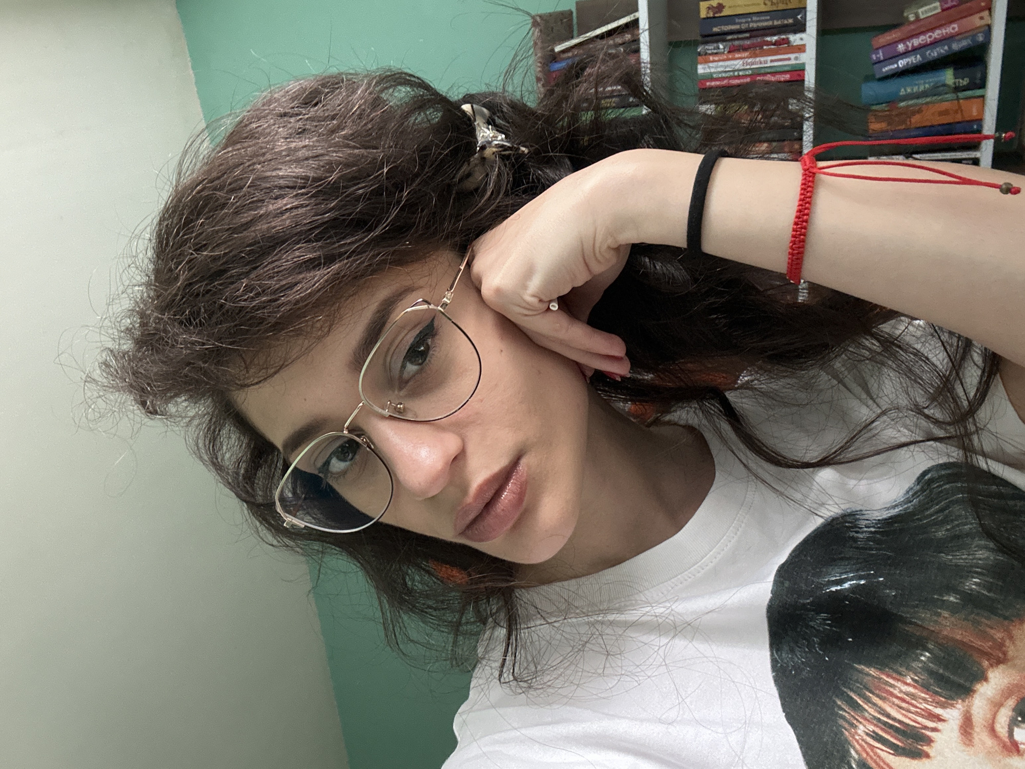 The media file is a close-up, selfie-style photograph of a young woman with dark, wavy hair. She is wearing gold-rimmed glasses and has on a white t-shirt with a graphic print of a person's face on it. Her right hand is resting on her face, near her eye, and she is wearing a red string bracelet on her wrist. In the background, there are bookshelves filled with books. The lighting suggests it is daytime. The woman's expression appears neutral or slightly contemplative. The location cues (bookshelves) and the general setting suggest an indoor environment, possibly a home or study. The overall mood is casual and personal. There is visible text on the spines of the books in the background, but it is mostly illegible at this resolution, though some appear to be in Cyrillic script. The image was likely taken in Sofia, Bulgaria.