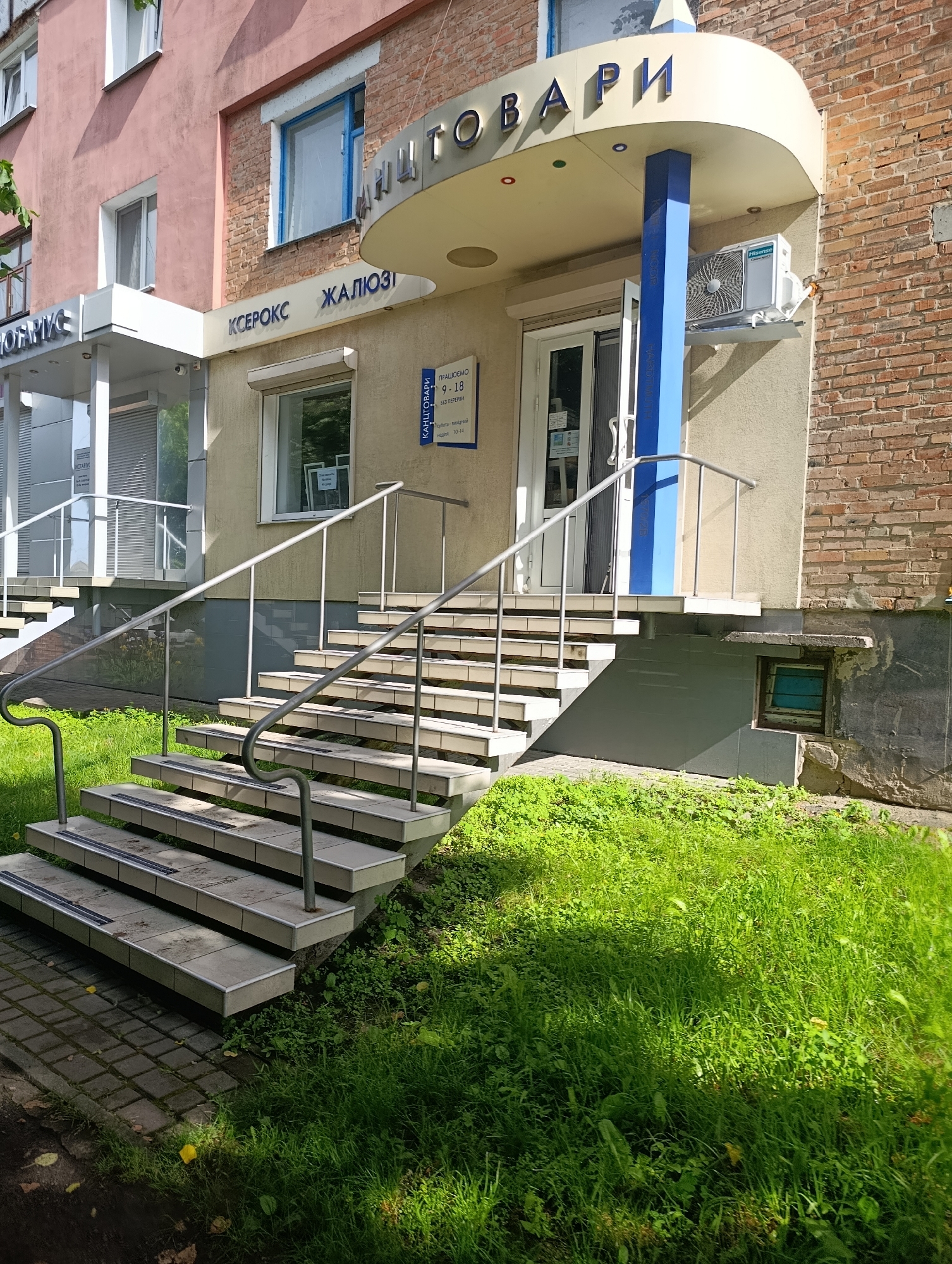 The image shows the exterior of a commercial building with a prominent entrance featuring a set of outdoor stairs and a covered walkway. The building has a pink facade on the left and a brick facade on the right. Above the entrance, there is a curved awning with blue lettering that reads "КАНЦТОВАРЫ" (meaning "stationery"). Below the awning, there is a sign that reads "Ксерокс Жалюзи" (meaning "Xerox Blinds"). A blue support column is visible in front of the entrance. An air conditioning unit is mounted on the wall to the right of the entrance. The stairs are made of concrete with gray tile treads and have metal handrails. The area around the stairs is covered with green grass. A paved walkway is visible at the bottom of the stairs. The image appears to have been taken during the daytime. The location is identified as Khorol, Ukraine.