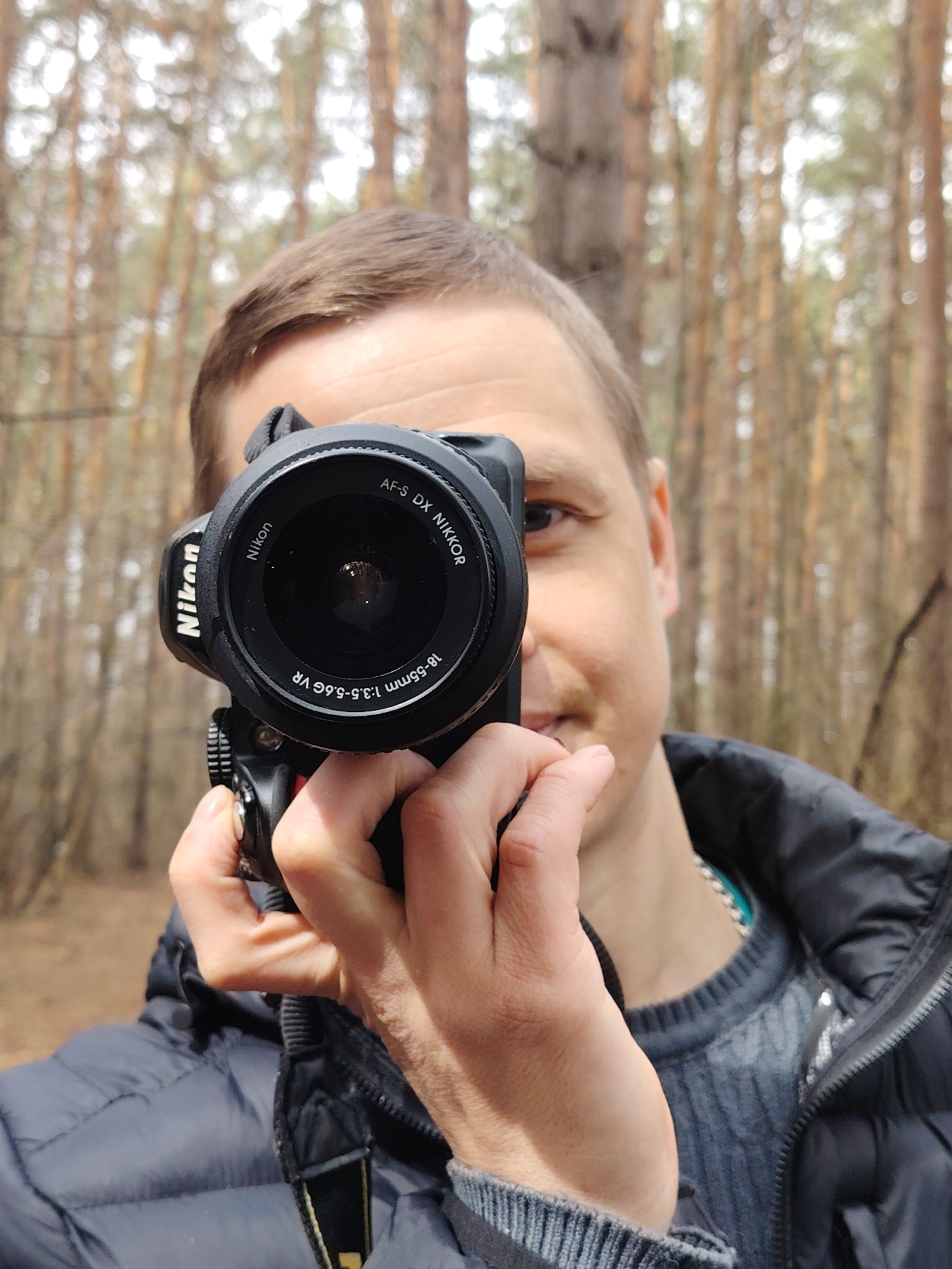 A close-up, waist-up shot depicts a Caucasian male holding a Nikon camera in front of him. He is wearing a dark puffy jacket over a gray sweater with a round neck. The background consists of a blurred forest of pine trees. The location is the city of Chaikovsky, Russia.