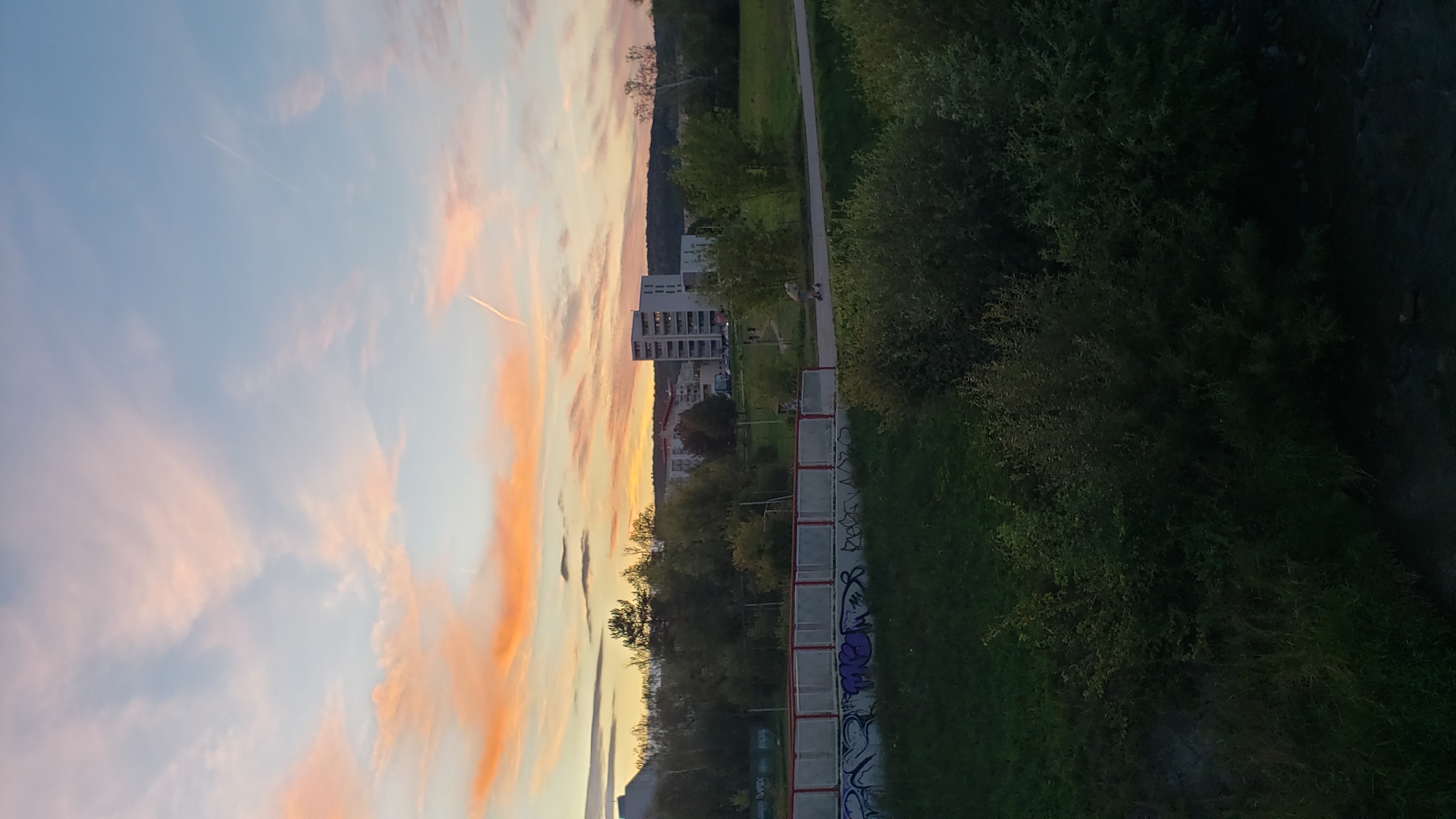 The image captures a sunset scene in the city of Prešov, Slovakia. The sky is filled with soft orange and pink hues, with scattered clouds and airplane contrails. In the mid-ground, there are buildings, including a tall, multi-story residential building. Trees and grassy areas are visible, suggesting a park or residential neighborhood. A red-railed bridge with graffiti on its lower wall spans across a grassy area in the foreground. A person on a scooter is visible on a paved path. The overall atmosphere is calm and serene, indicative of early evening.