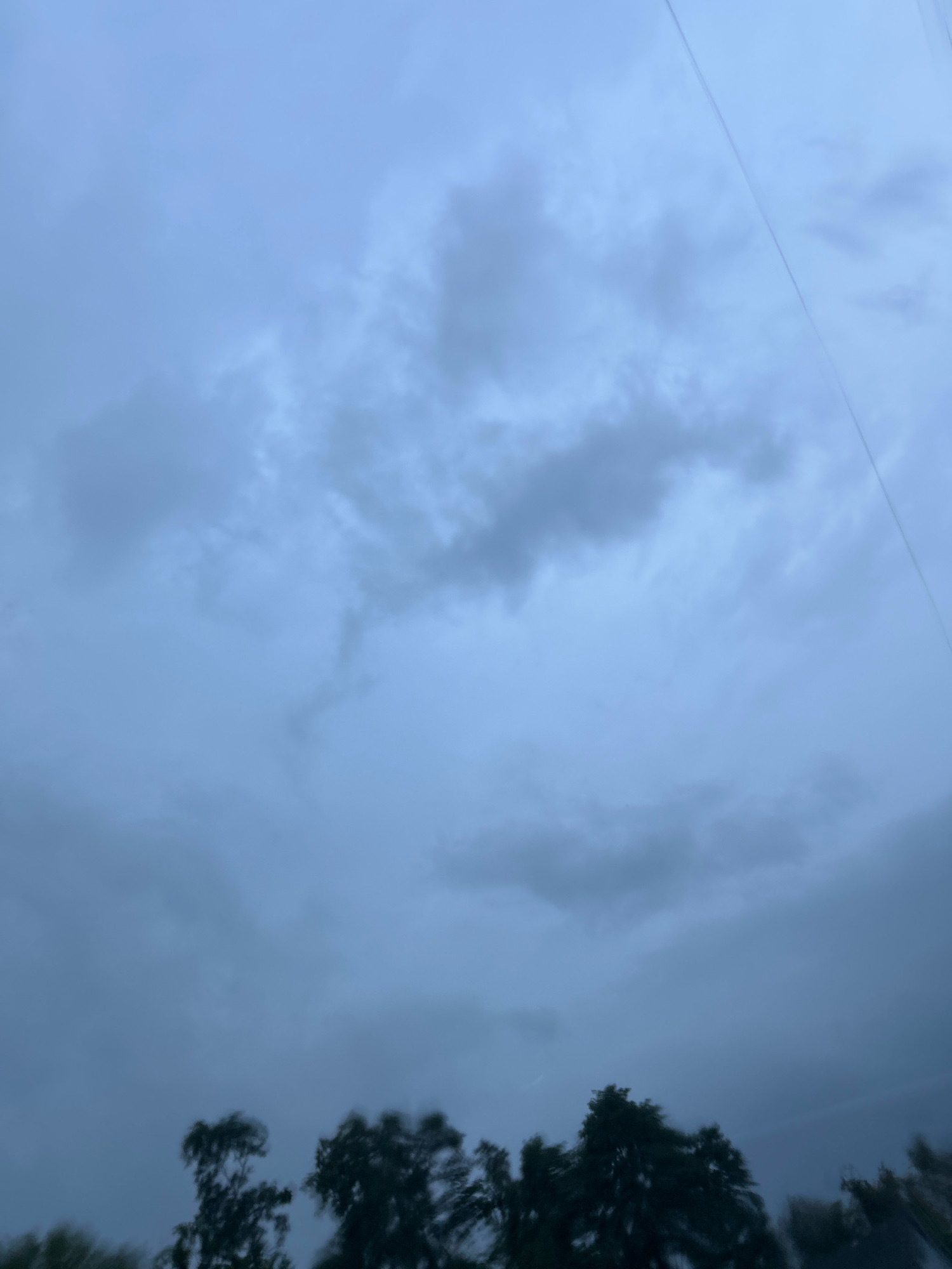 The image shows a cloudy sky with a few trees visible at the bottom. The sky is a muted blue-gray, with various shades of clouds creating a textured effect. A thin wire runs diagonally across the upper right portion of the frame. The trees at the bottom are silhouetted against the sky, appearing dark and slightly blurred, suggesting motion or low light conditions. The overall impression is of an overcast day. The location is identified as Scott Township, United States of America.