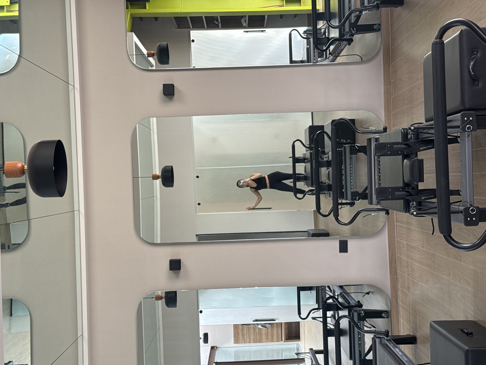The image depicts the interior of a modern fitness studio, likely a Pilates studio, characterized by light-colored walls and numerous mirrors with rounded corners. The photo is oriented ninety degrees counter-clockwise, making the walls appear as the floor and ceiling.
The primary subject is a woman, reflected in the central mirror. She is wearing a black sports bra or crop top and black leggings. She is captured in the act of performing an exercise, appearing to be a side plank or similar core strengthening move, supported by one hand and her feet on a Pilates reformer. With her other hand, she is holding a smartphone up to her face, suggesting she is taking a mirror selfie or recording her workout.
Several black Pilates reformers are visible, both directly in the foreground and reflected in the mirrors. One piece of equipment clearly shows the brand name "Balanced Body". Black, cylindrical lighting fixtures or decorative wall accents with wooden bases are mounted on the walls and also visible in reflection. Small, black square objects, possibly electrical outlets or switches, are also present on the wall. The reflections in the upper and lower mirrors hint at more of the studio's interior, including a bright yellow structure overhead and what appears to be wooden shelving in the background. The flooring in the direct view (right side of the image) is light-colored wood or wood-patterned tile. The studio appears well-lit and clean.