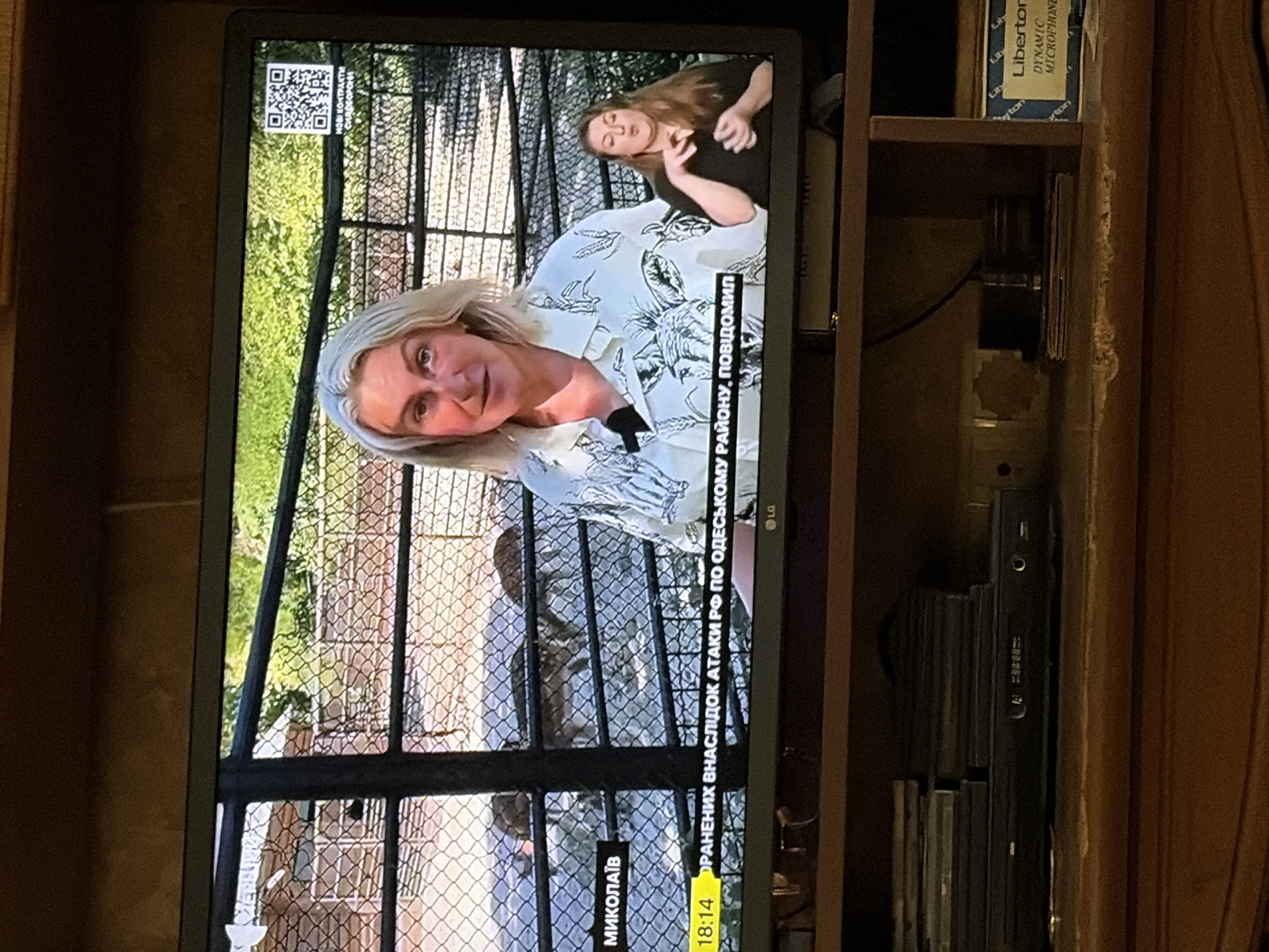 The image shows a television screen displaying a news broadcast. On the screen, a blonde woman is speaking, likely a reporter or news anchor. She is wearing a patterned shirt. To her right, a woman is signing, presumably providing sign language interpretation for the broadcast. Behind them, a fenced enclosure with some deer can be seen. A ticker at the bottom of the screen shows "18:14" and text in a foreign language. A QR code is also visible in the upper right corner of the screen. Below the television, a DVD player and a stack of discs are visible on a shelf. Further down, another shelf holds a box labeled "Liberton DYNAMIC MICROPHONE". The background appears to be a wooden cabinet.