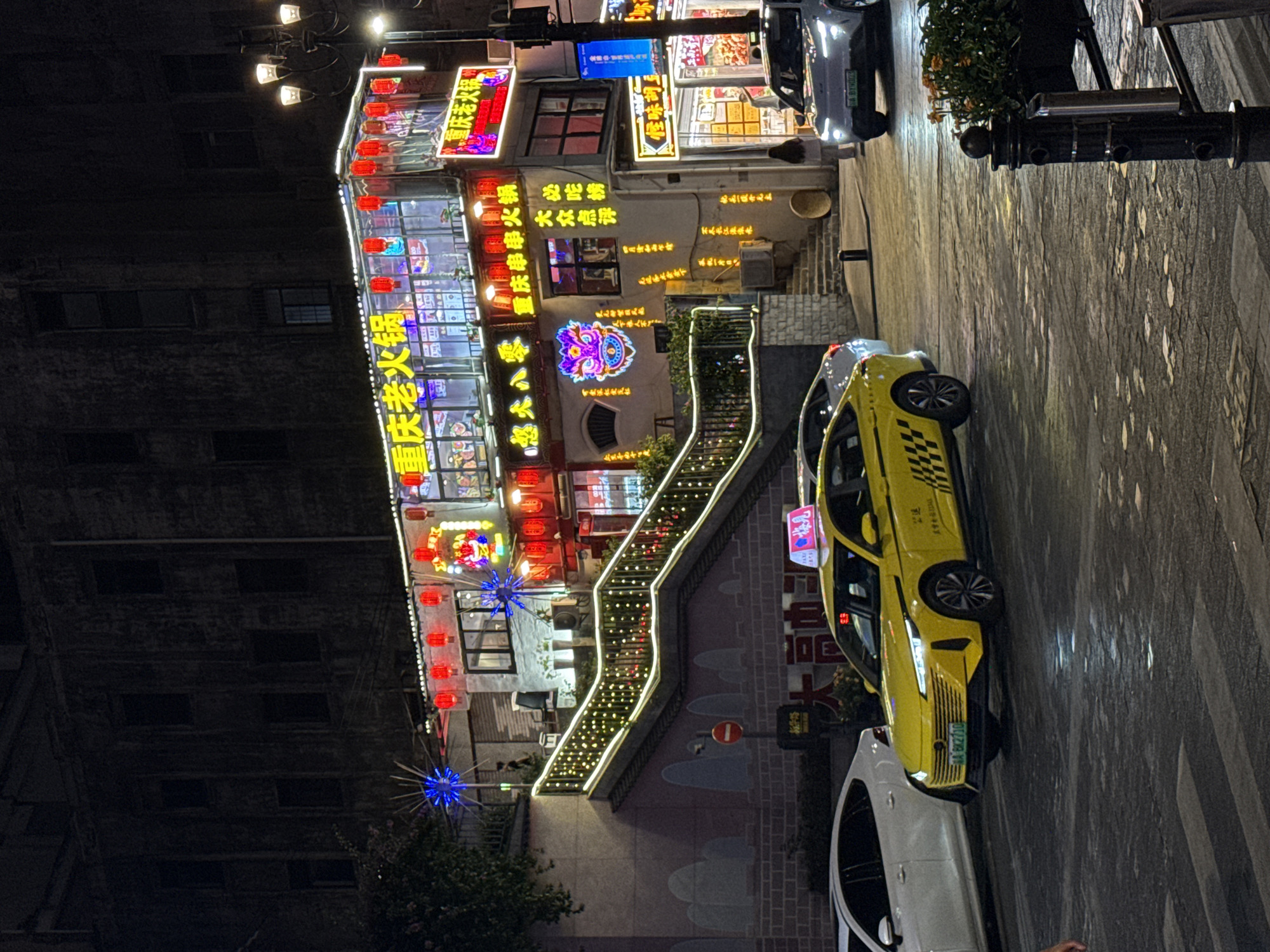A night view of a busy street in Yuzhong District, China, showcases a vibrant restaurant illuminated by numerous red lanterns and neon signs. The building features multiple stories adorned with bright signage in Chinese characters, hinting at a lively dining establishment, likely a hot pot restaurant given the prominent text. A set of stairs, also lit with string lights, ascends to the restaurant's entrance, guiding patrons up from the street level.
In the foreground, a bright yellow taxi with checkered patterns on its side is parked or slowly moving on the wet pavement. A white car is visible behind it. Further down the street, another vehicle, a light-colored car, is seen approaching. The overall atmosphere is bustling and energetic, typical of a commercial district at night. The architecture suggests a mix of traditional and modern elements, common in Chinese urban landscapes. The street appears to be made of cobblestones or a similar textured material. The presence of streetlights and the illuminated building create a warm, inviting glow against the dark night sky.