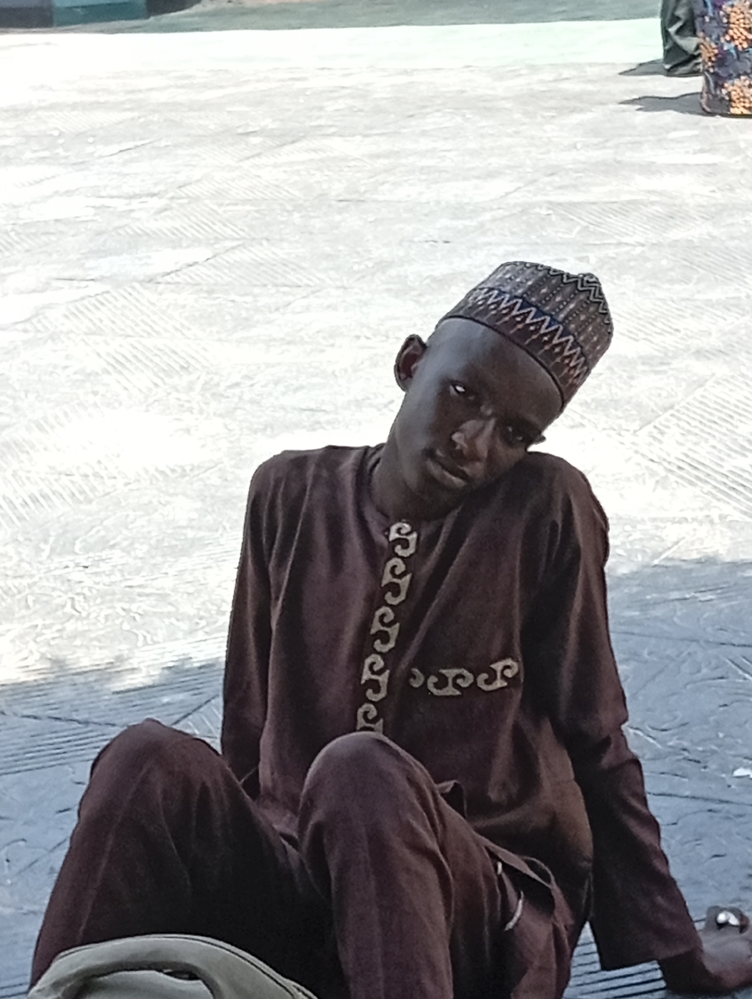 A young Black man is sitting on a tiled surface. He is wearing a dark brown, long-sleeved shirt with light-colored decorative embroidery along the front and a dark brown cap with a lighter colored patterned brim. He is looking at the camera with his head tilted slightly to the side. The tiled surface has a slightly textured pattern. A small portion of a person's leg and clothing are visible in the upper right corner. The location is Dadi Riba, Nigeria.