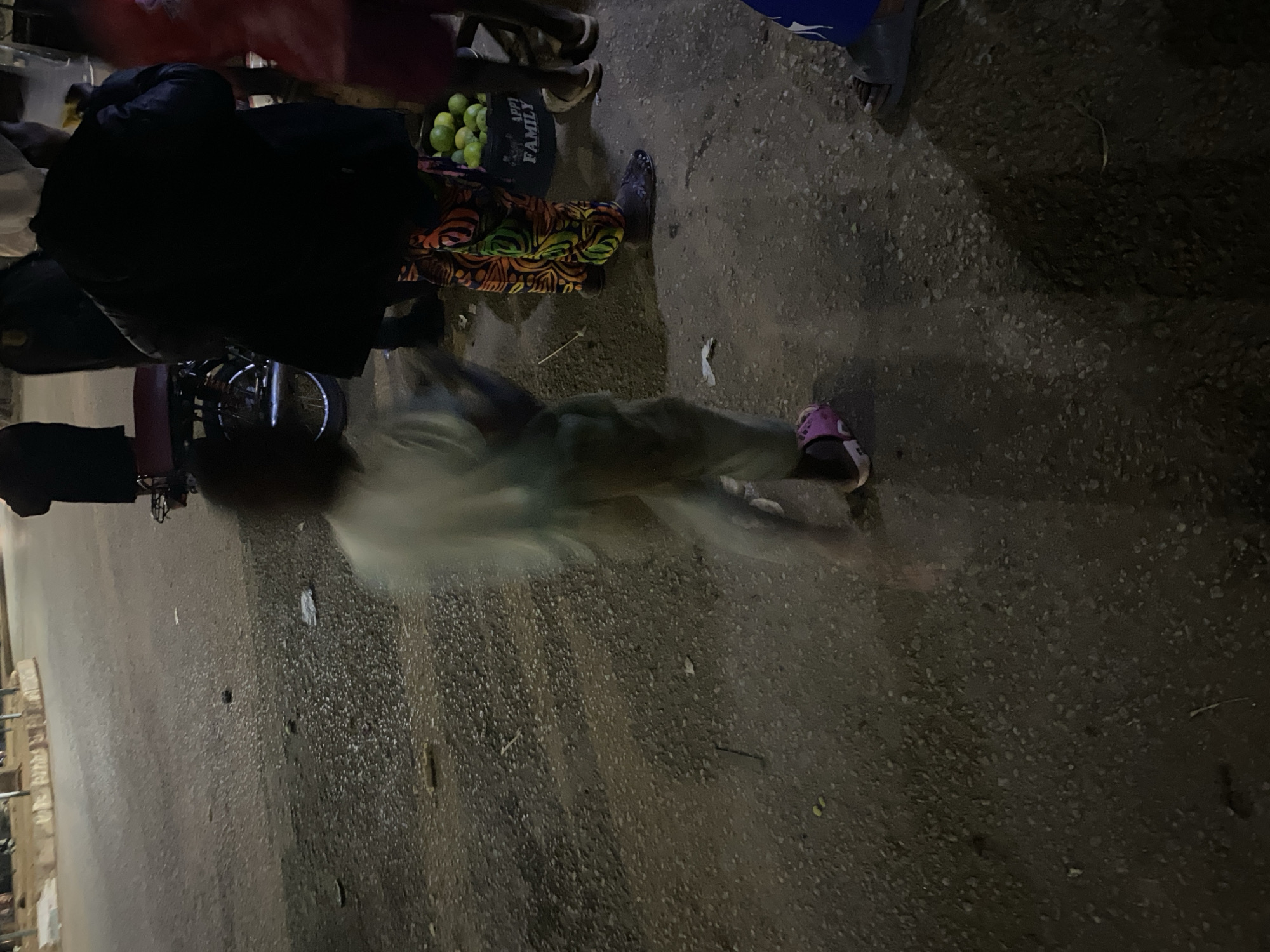 The image captures a scene on an asphalt road in Bukuru, Nigeria, at night. Several individuals are present, some partially obscured by motion blur due to the low light conditions. In the foreground, a child wearing a light-colored garment and pink shoes is in motion, creating a blurred effect. To the right, an adult wearing a dark jacket and vibrantly patterned trousers stands next to a black bucket filled with green produce, likely limes or lemons, with the word "FAMILY" visible on the bucket. Another person in a red garment is partially visible on the far right. In the background, a motorcycle is parked, and other figures are faintly discernible, suggesting a public space or marketplace. The ground is rough and gritty, consistent with a road surface, with scattered debris. The lighting is dim, indicating it is nighttime or a poorly lit area.