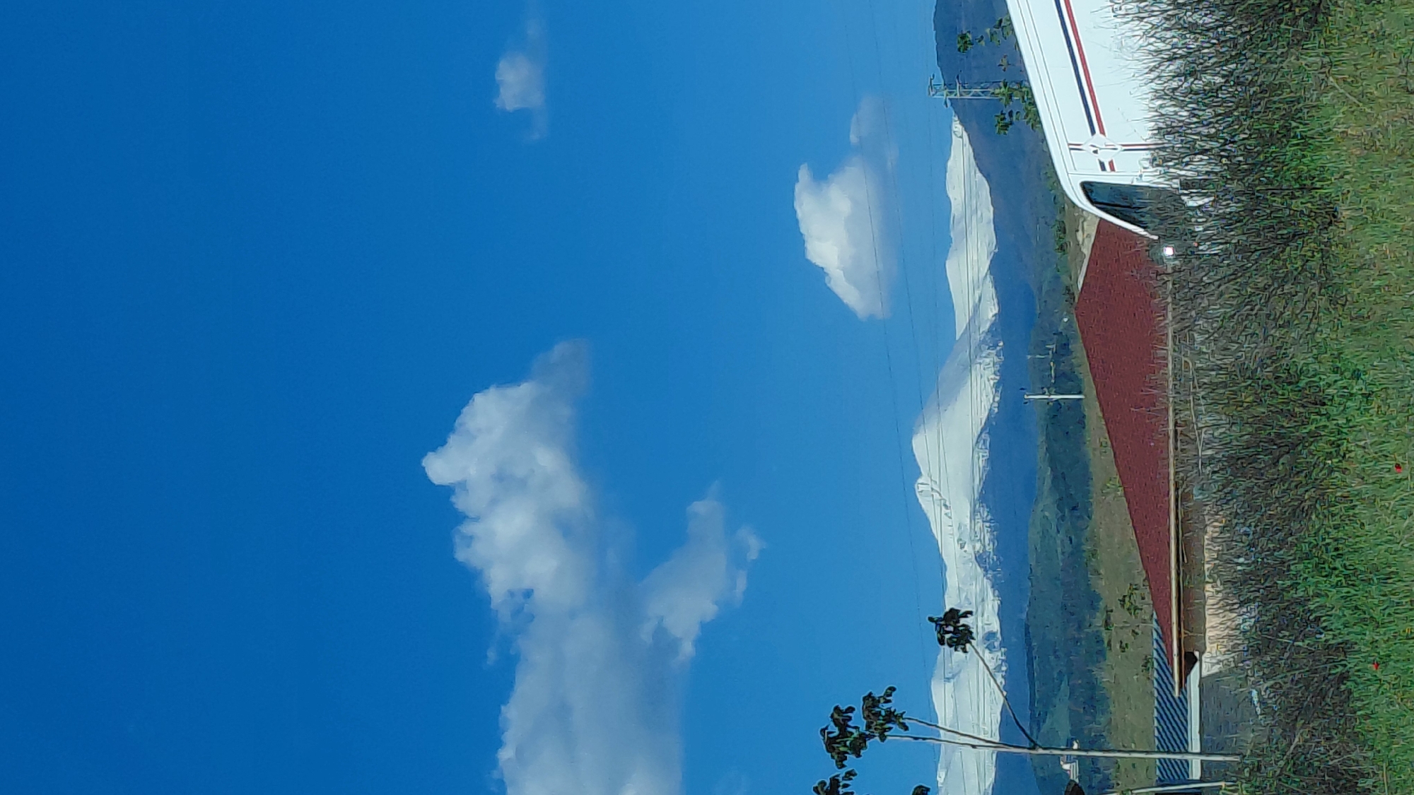 The image shows a landscape with a bright blue sky dotted with puffy white clouds. A range of snow-capped mountains is visible in the background. In the foreground, there's a grassy area with bushes and a partially visible building or structure featuring a red roof and white siding. A white vehicle with red and blue stripes is parked next to the structure. A few trees are also present. Power lines are visible against the mountains. The location is likely a rural area with scenic views.