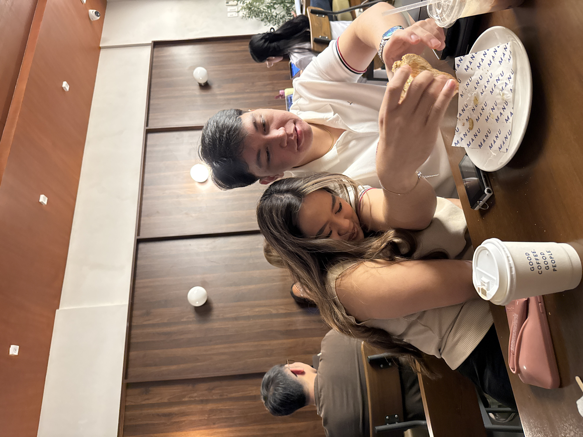 Here is a concise description of the image:
The photo shows a man and a woman seated closely at a table in what appears to be a cafe or restaurant. The man is holding and presumably eating a sandwich over a plate lined with MANU brand paper, while the woman rests her head affectionately on his shoulder. A coffee cup is beside the woman, its label reading "GOOD COFFEE. GOOD PEOPLE." The scene is well-lit, suggesting an indoor environment with modern decor, including a wood-paneled wall. The setting is in Davao City, Philippines, based on the MANU brand mentioned on the plate.