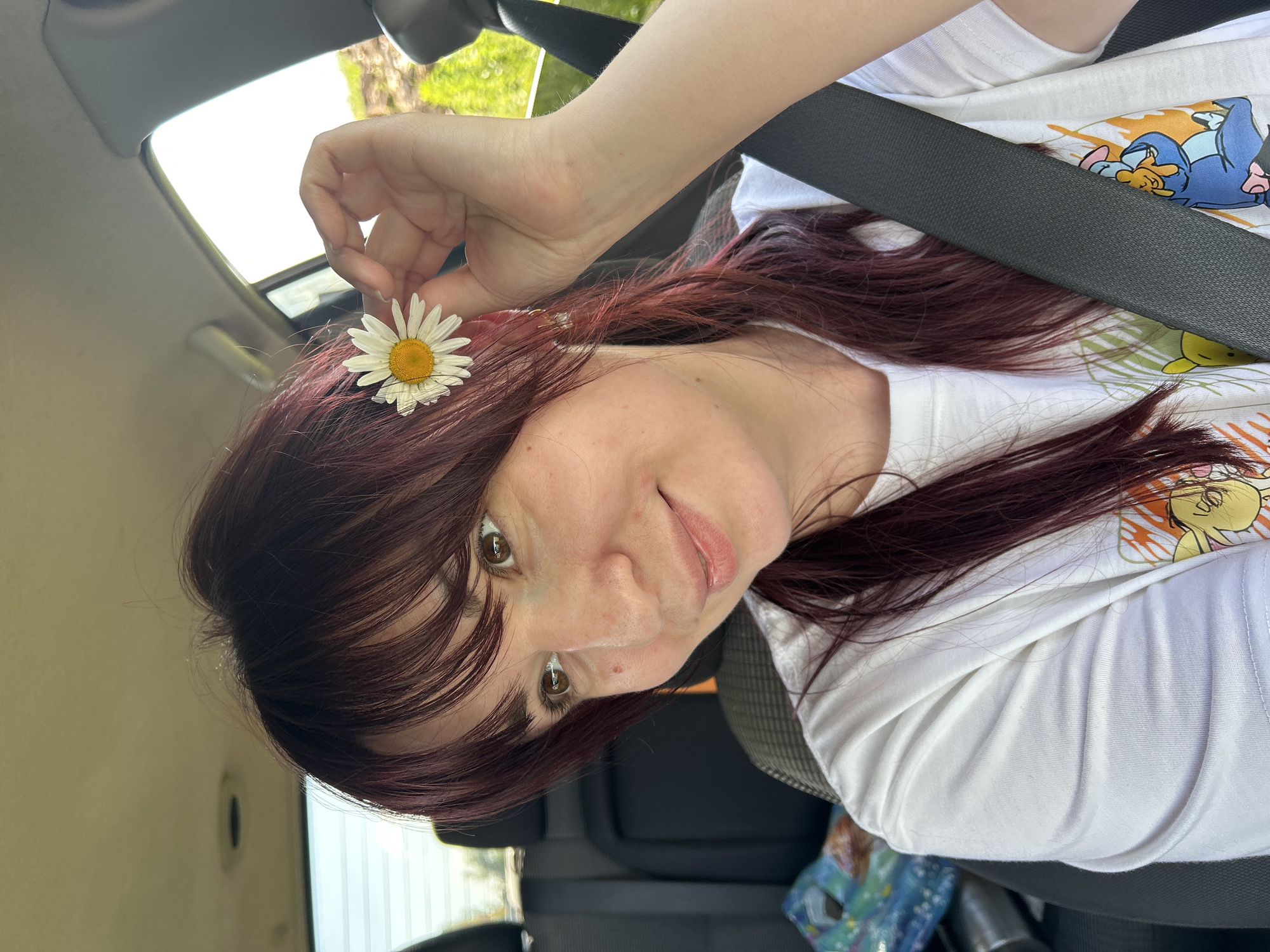 A woman with dark red hair is in the passenger seat of a vehicle, holding a daisy to her hair. She is wearing a white t-shirt with a cartoon graphic and a seatbelt. The interior of the car is visible, as is a blurred outdoor scene through the windows. The image was taken in the city of Fokino, Russia.