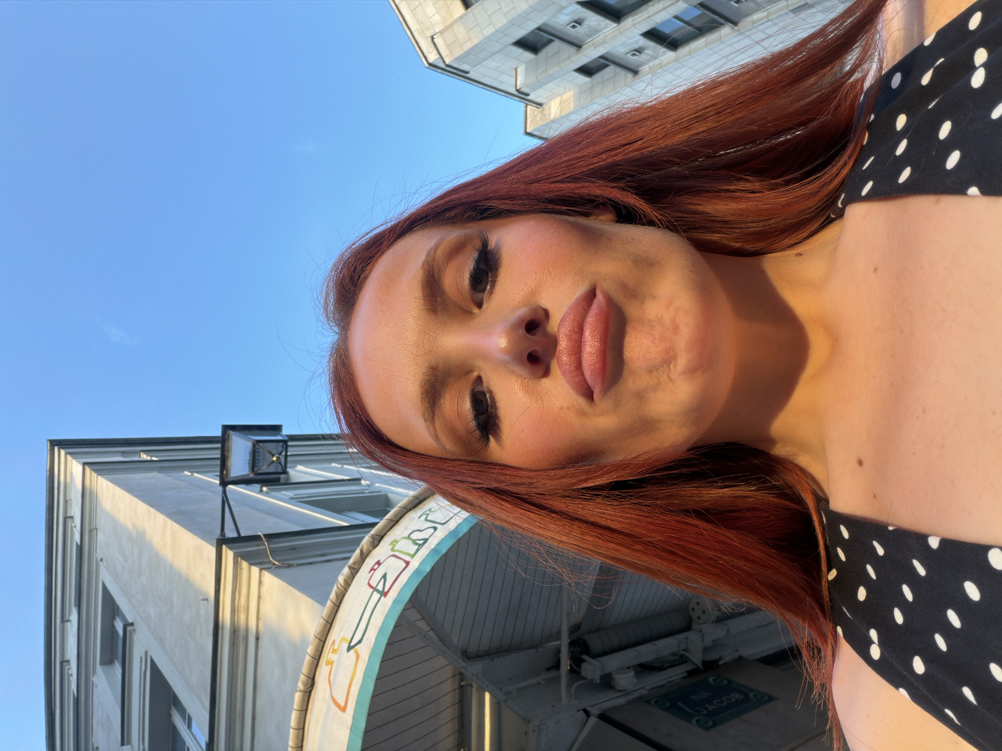 A downward-angled, medium close-up shot depicts a fair-skinned woman with red hair, focusing primarily on her head and upper torso, set against an urban backdrop. She is wearing a black and white polka-dotted top.
Behind her, buildings rise against a clear blue sky. One building is light-colored with evenly spaced windows and a lamppost affixed to its upper corner. The other building in the upper right portion of the frame shows repeating windows on its multiple floors.
Below the woman, an archway is visible, featuring a decorative border with repeated drawings of objects. Beneath the archway, the surface has a sign that reads “RUE JACOB” and an arrow.
The presence of "RUE JACOB" indicates that the location is in Paris, France.