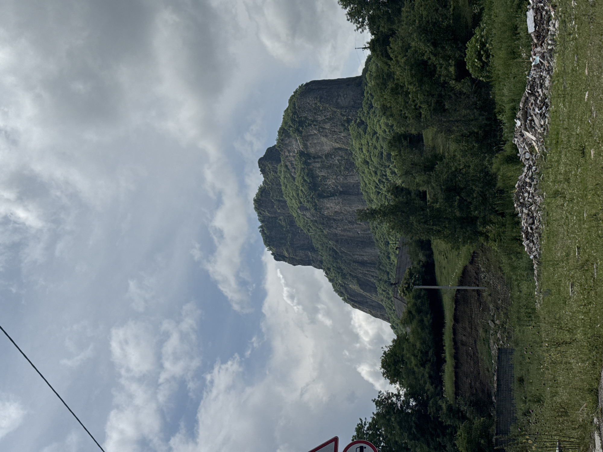 A tall, rugged mountain with a jagged peak dominates the center of the frame, its steep slopes covered in dense green vegetation. The mountain is set against a sky filled with dramatic, billowing clouds, casting shadows that accentuate the mountain's textures. In the foreground, lush green grass and wildflowers grow, interspersed with a pile of rubble and building debris. To the left, a traffic sign with a red border and a white background is visible, along with a portion of a dark pole. A thin, dark power line stretches diagonally across the upper left portion of the image. Trees line the base of the mountain and extend to the sides, with a building or structure with a dark, pitched roof visible nestled among them. The overall impression is a natural landscape with hints of human presence.
