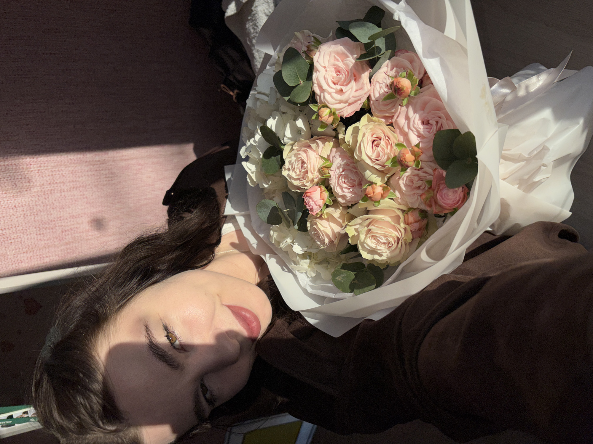 A young woman is captured in a close-up portrait, lying down next to a large bouquet of flowers. She occupies the left side of the frame, looking directly at the viewer with a gentle, subtle smile. Her dark brown hair cascades around her face, and she wears a dark brown top or garment. A striking diagonal shadow falls across her face, emphasizing her warm brown eyes and soft features.
Beside her, filling the right side of the image, is a lush bouquet of flowers. It features numerous pale pink and cream-colored roses, some tightly budded and others in full bloom. White hydrangea clusters and round green eucalyptus leaves are interspersed among the roses, adding volume and texture. The bouquet is elegantly wrapped in white paper, secured with a delicate white ribbon, suggesting it's freshly received.
The scene appears to be indoors, bathed in bright, natural sunlight, which casts distinct shadows across the woman's face and the background, indicating it's likely morning or late afternoon. A soft, light pink textured surface, possibly a carpet or blanket, is visible in the background, contrasting with a darker, indistinct area. The overall atmosphere is serene and joyful, highlighting the beauty of the flowers and the woman's pleasant expression. No discernible text is visible within the image. The location context provided is Perm, Russia.