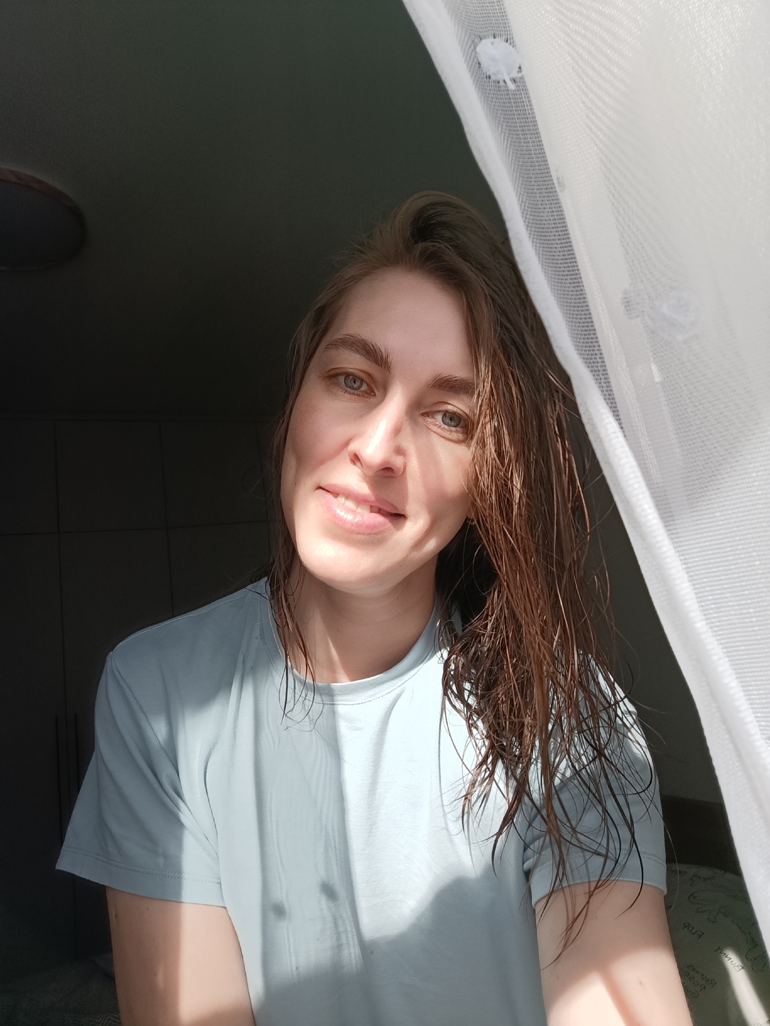 A woman poses for a selfie. She is wearing a light blue t-shirt and has wet, brown hair. She is smiling slightly. A sheer, white curtain is on the right side of the frame. In the background to the left, there is a dark room with a cabinet. The location is Chaikovsky, Russia.