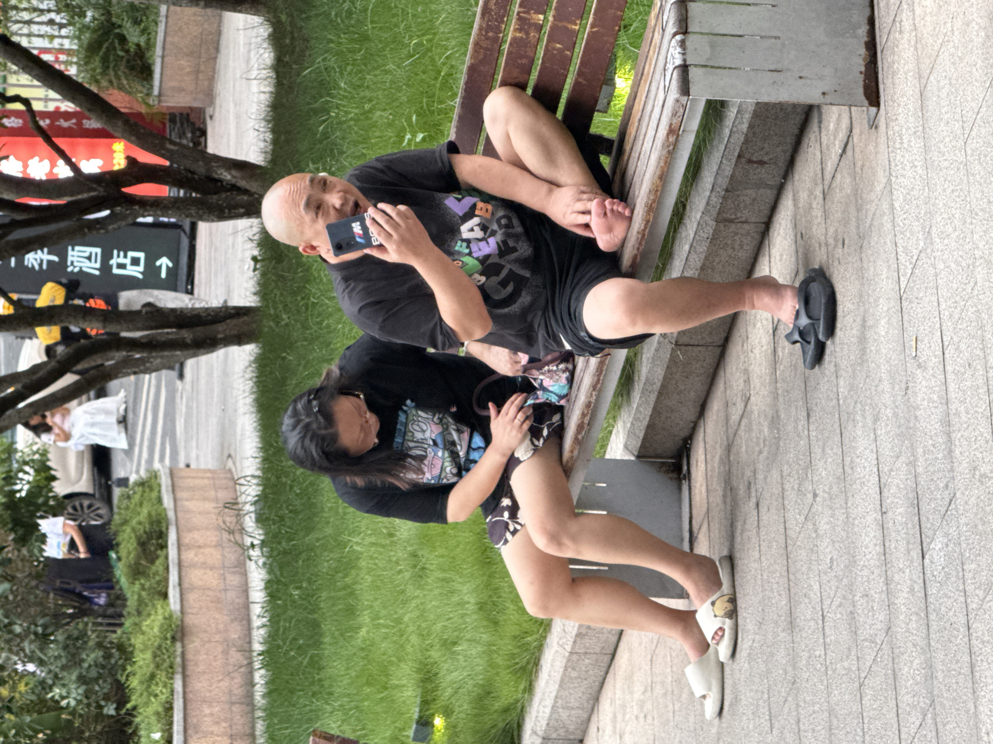In the city of Yuzhong District, China, a man and a woman are sitting on a wooden bench. The man, who is bald, is holding a smartphone and looking at it with his mouth open. He is wearing a black t-shirt with graphics on it and black shorts. The woman, sitting to his left, is wearing glasses and a black t-shirt with a graphic print. She is wearing a patterned skirt and white slippers. Her left hand is resting on a bag. The bench is situated on a paved area, with a grassy embankment behind it. In the background, there are trees, a sign with Chinese characters and an arrow, and other people in the distance, including a woman in a white dress.