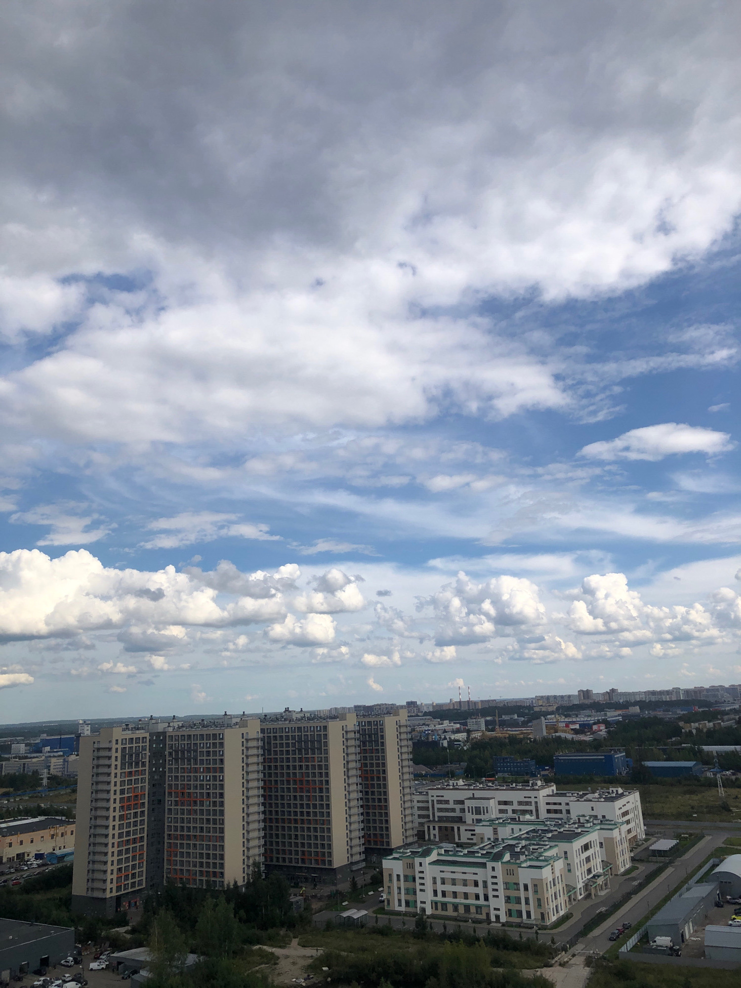 An aerial, wide-angle shot captures a sprawling urban landscape under a dynamic, cloudy sky in an unknown city in Russia. In the foreground, a cluster of tall, modern residential buildings with beige and gray facades and numerous windows are prominent. To the right, a complex of lower-rise, white and beige apartment buildings is visible, featuring a modern architectural style with multiple interconnected sections. The midground is filled with more buildings, industrial structures, and green spaces, with chimneys visible in the distance. The background shows a hazy horizon of more urban development. The sky is a mix of light blue and varying shades of gray clouds, with dramatic streaks and formations. The overall scene depicts a contemporary residential and industrial area with a vast sky overhead.
