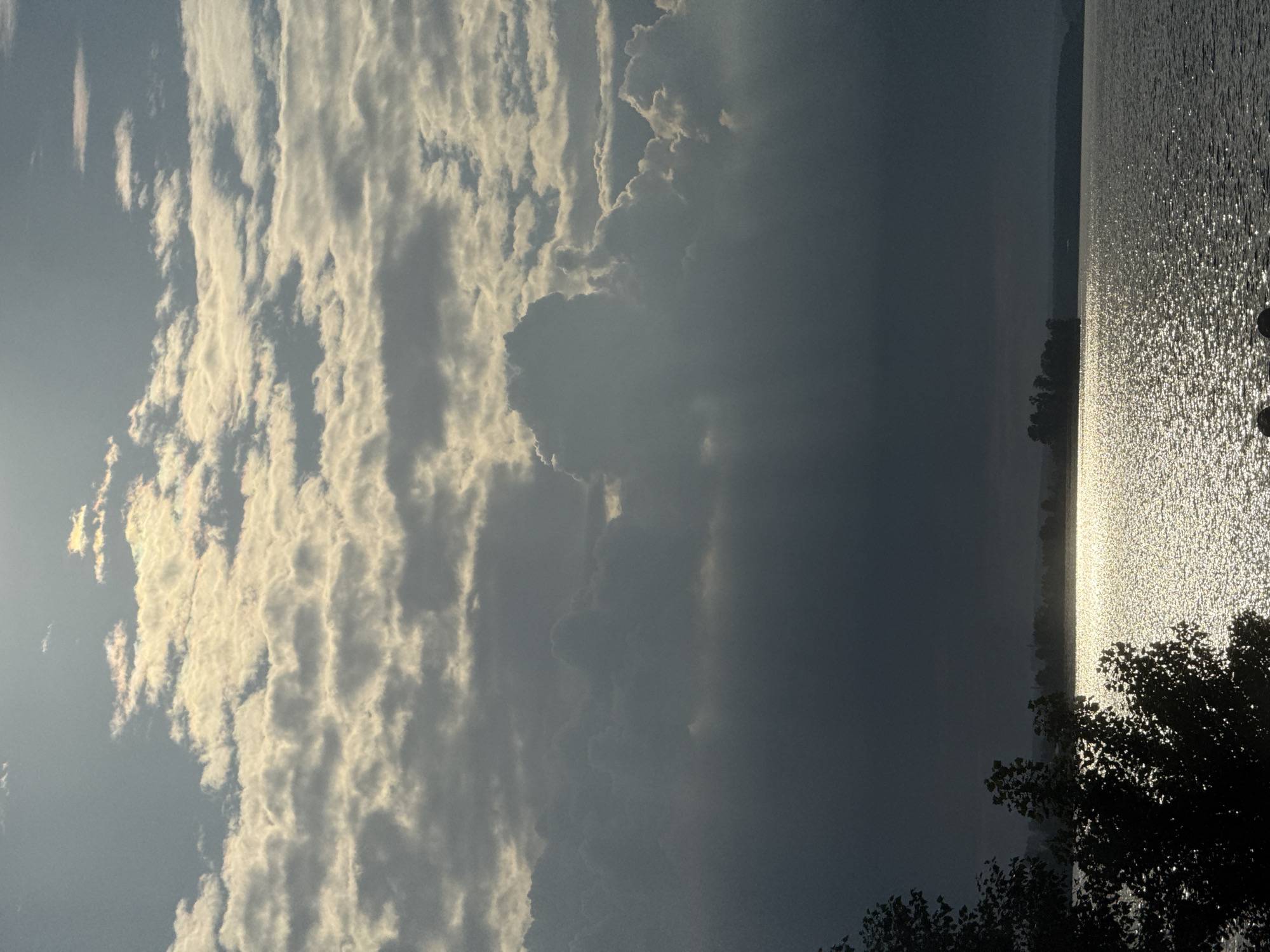 A body of water, likely a lake or river, reflects the light of the sun, which is partially obscured by dramatic, layered clouds. The clouds range in color from bright white where the sun hits them to dark gray where they are shadowed. The water's surface is covered in a shimmering pattern of light reflections. In the foreground, the dark silhouette of trees and foliage is visible on the left side of the image. In the distance, a dark, silhouetted shoreline with a few scattered trees can be seen. The overall impression is of a dramatic, possibly stormy or post-storm, sky over a body of water. The location is Votkinsk, Russia.