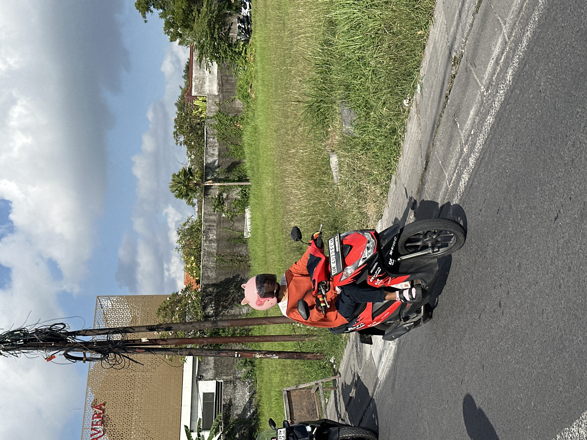 In North Kuta, Indonesia, a person wearing an orange long-sleeved shirt and dark pants rides a red and black scooter on a paved road. They are wearing a pink helmet shaped like an animal's head with ears. The scooter has a license plate that reads "DK 4022 FCH". Another black scooter is parked to the left of the frame. In the background, there is a field of tall green grass, a concrete wall, and a modern building with a patterned facade and the word "VERA" visible. A utility pole with many tangled wires stands near the road. The sky is blue with scattered white clouds.