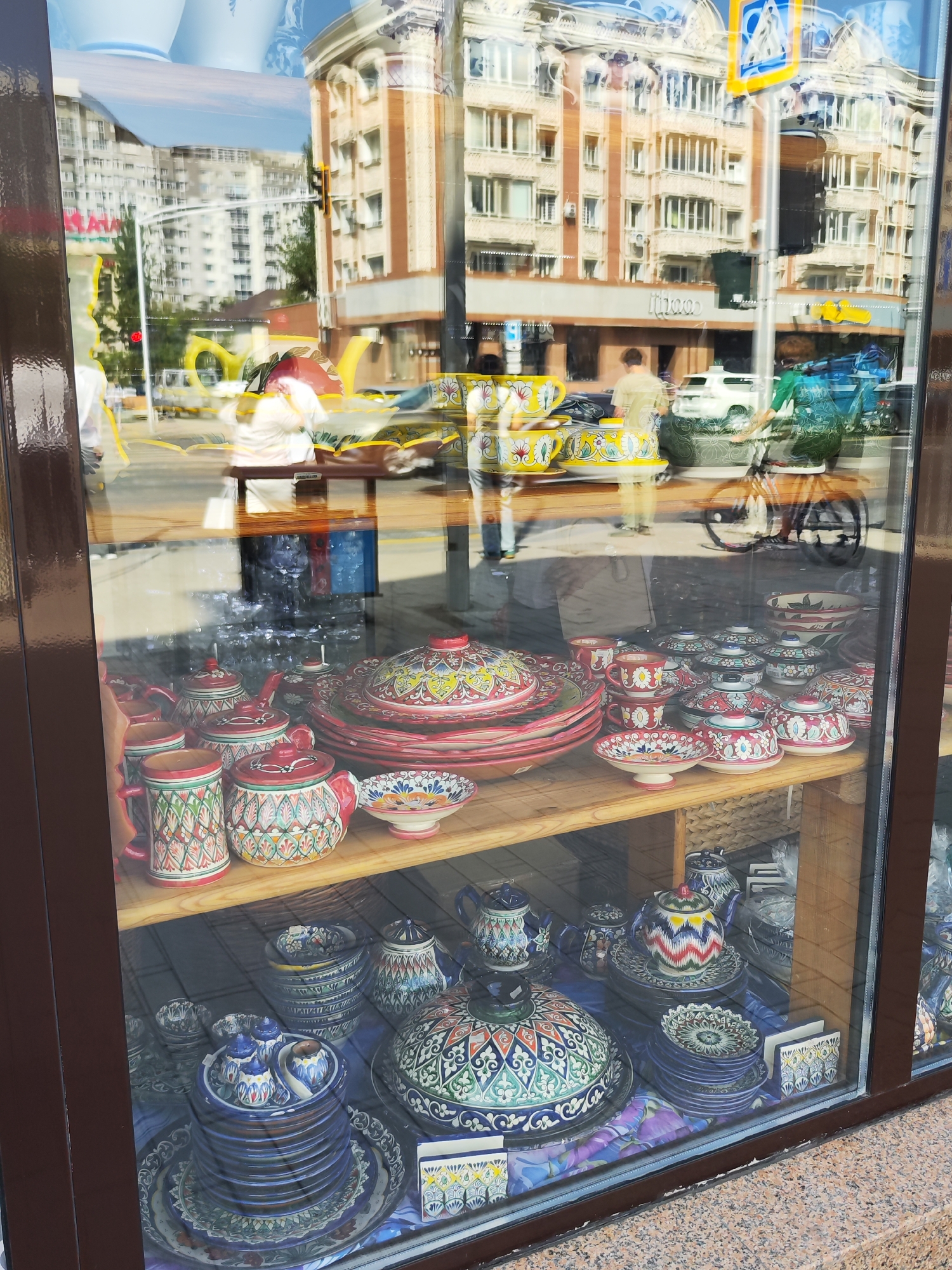 The image shows a reflection in a shop window in Astana, Kazakhstan. The reflection displays a street scene with buildings, traffic lights, people, and vehicles. In the foreground, inside the shop, there are shelves filled with decorative ceramic ware. The ceramics feature intricate, colorful patterns, with many pieces being teapots, bowls, and plates. The reflections on the glass create a layered effect, making it difficult to distinguish between what is inside the shop and what is reflected from outside.
